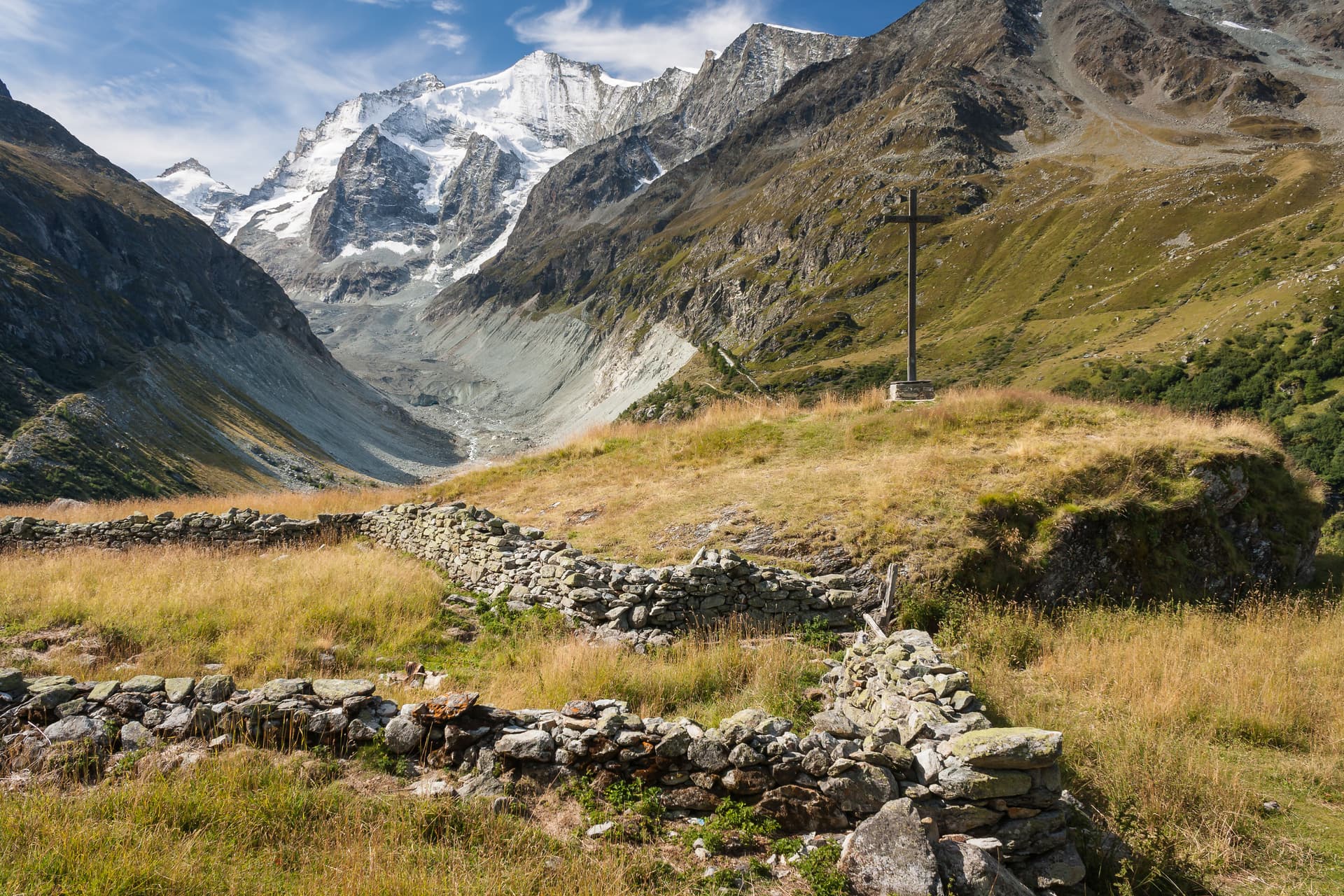 Alpine valley with snow-capped mountains, wooden cross, and stone wall in foreground.