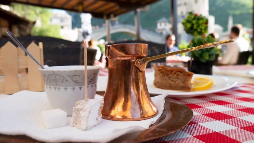 Bosnian coffee service with cezve, Turkish delight, and pastry outdoors overlooking a stone bridge.