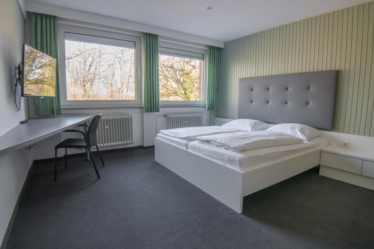 Modern hotel room with white bed, desk, and view of bare trees outside windows.