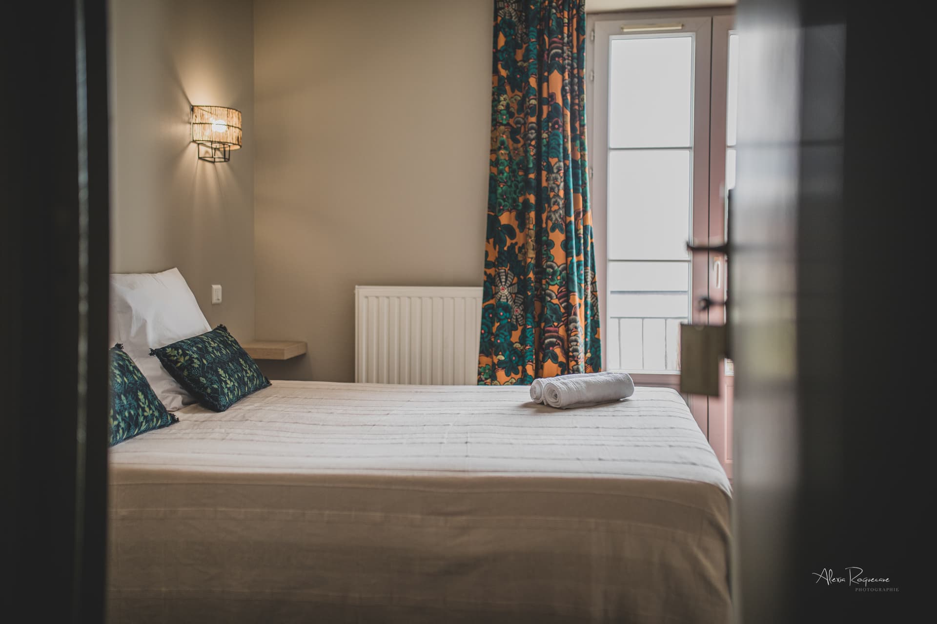 Bedroom with neatly made bed, patterned pillows, and bright floral curtains near a window.