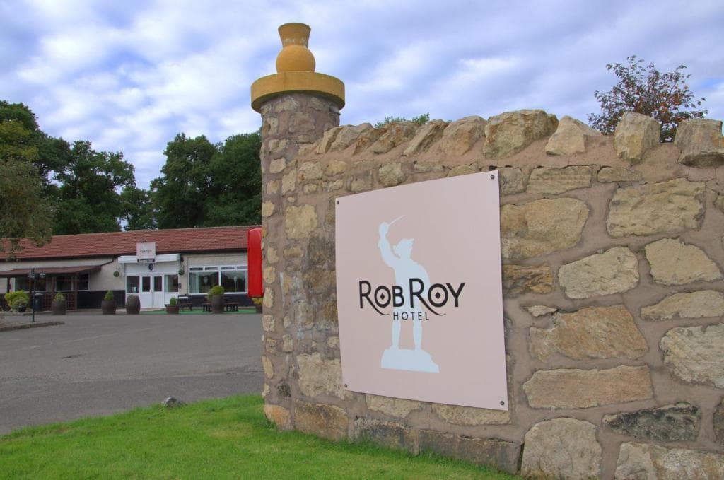 Rob Roy Hotel sign on stone wall with parking lot and low building in background