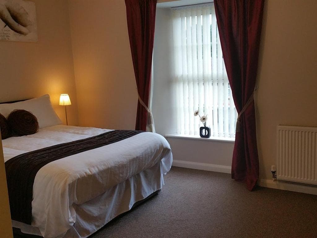 Bedroom with double bed, bedside lamp, window with vertical blinds, and red curtains at The Begelly Arms Hotel.