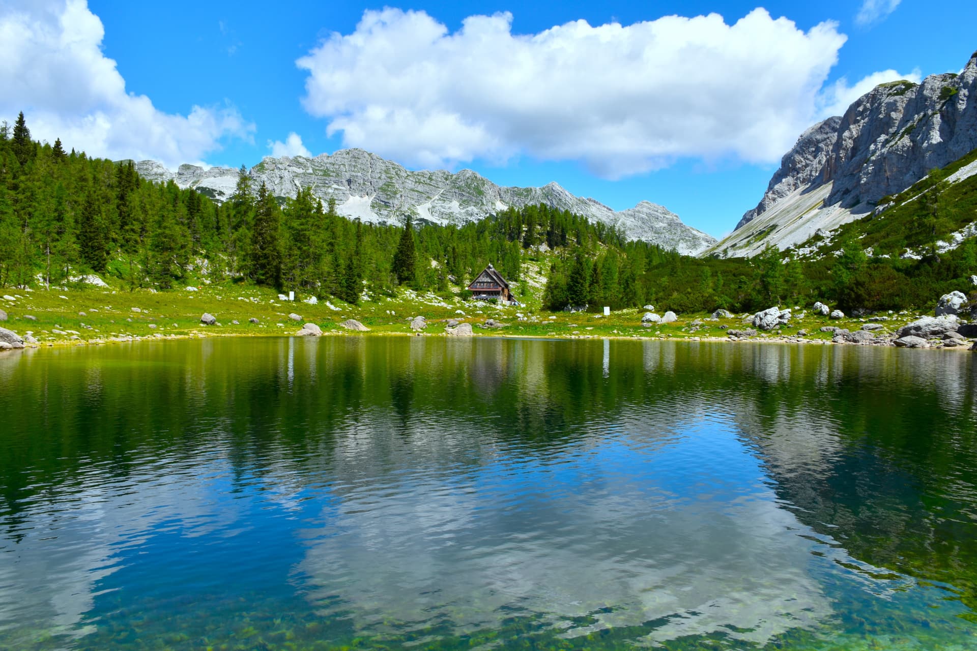 Alpine lake reflecting mountains and blue sky with clouds, small cabin visible.