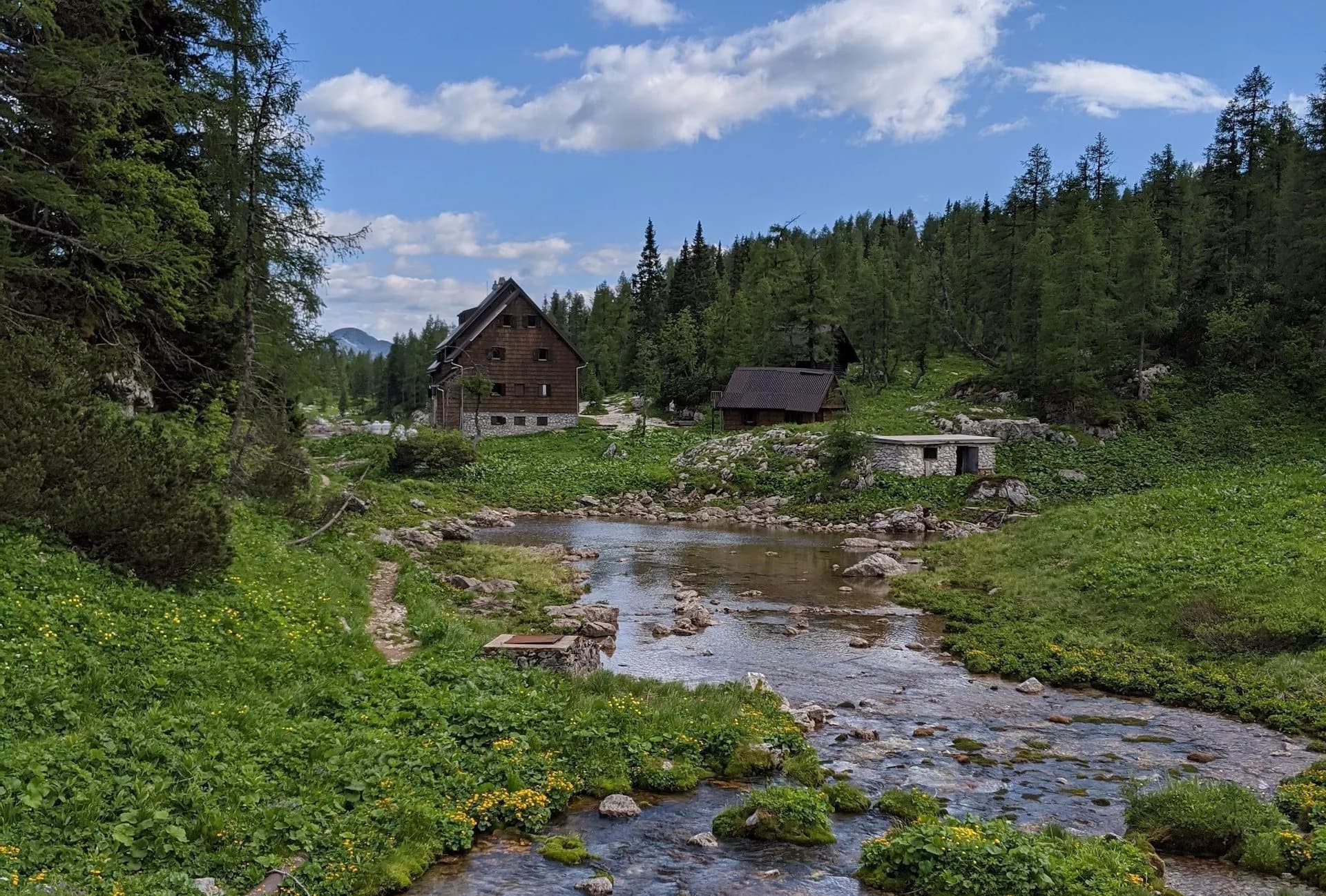 Mountain huts by a stream in a green valley under a blue, cloudy sky, Triglav Lakes.