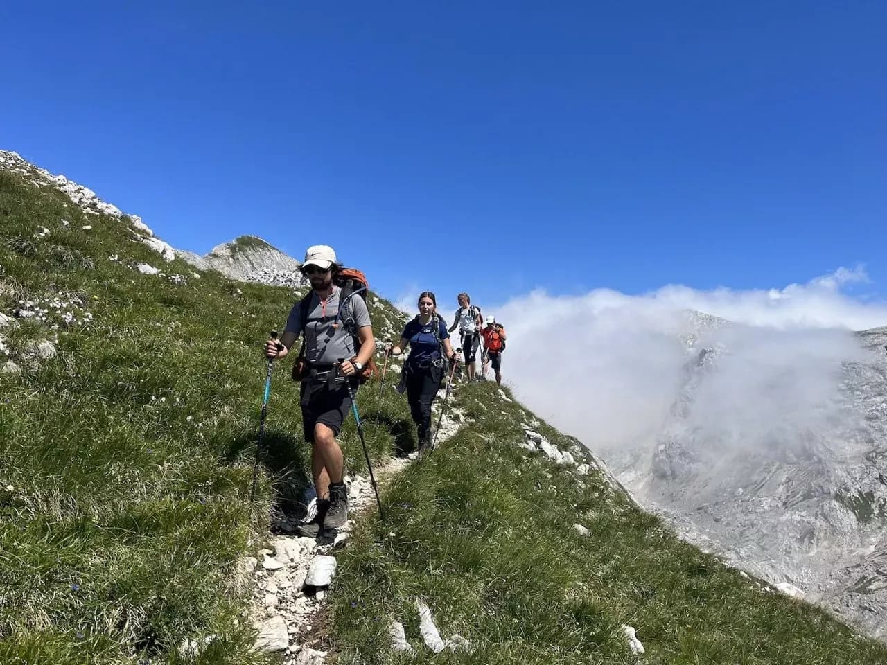Hikers with trekking poles traverse a narrow, grassy ridge above a valley with clouds below.