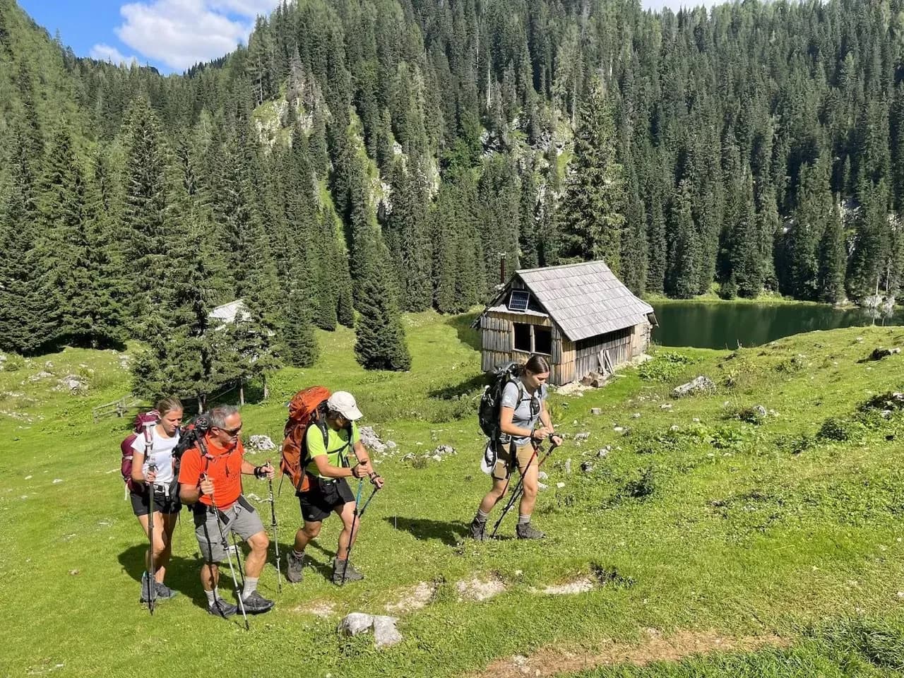 Hikers with backpacks and poles trekking past a wooden cabin near a mountain lake.