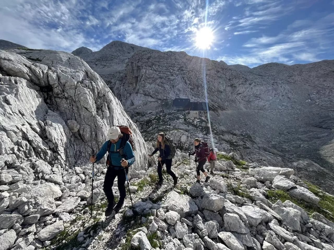 Hikers with backpacks ascend rocky terrain toward a mountain hut under a bright sun.