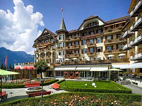 Grand hotel exterior with "Lauberhorn" sign, garden terrace, and mountains under a blue sky.