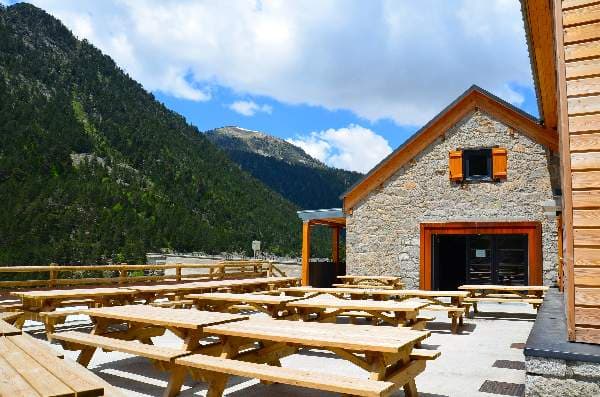 Picnic tables outside stone refuge with forested mountains at Refuge Lac de l’Oule