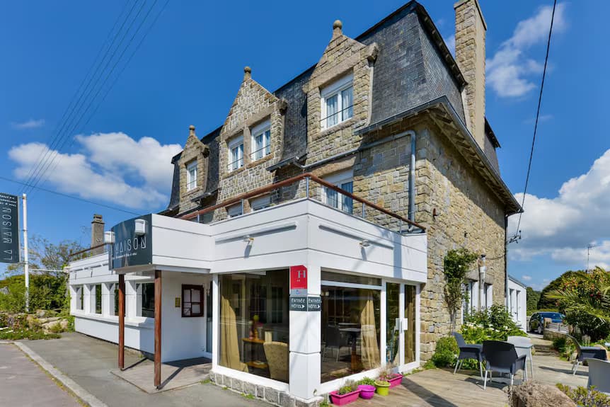 Stone hotel building with a white glass-fronted entrance and outdoor terrace seating under a blue sky.