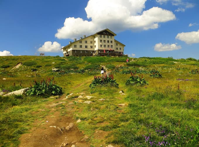 Hikers approaching Rila Lakes Hut on a grassy mountain slope under a blue sky.