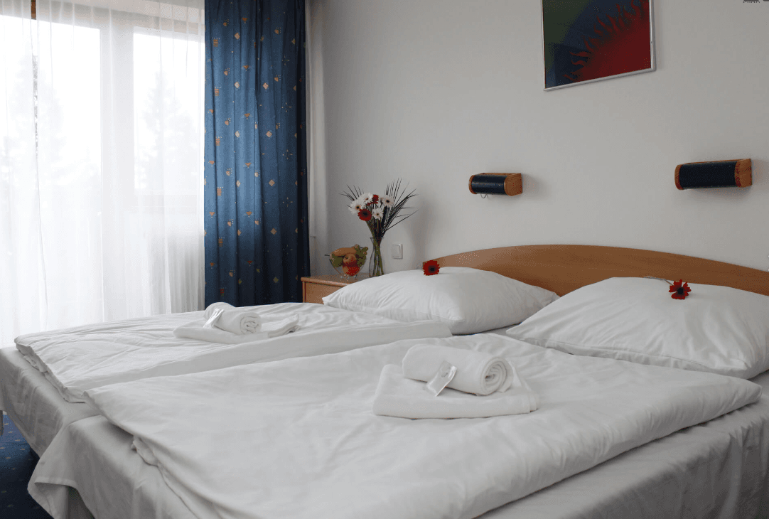 Twin beds with crisp white linens, rolled towels, and flowers in a bright hotel room.