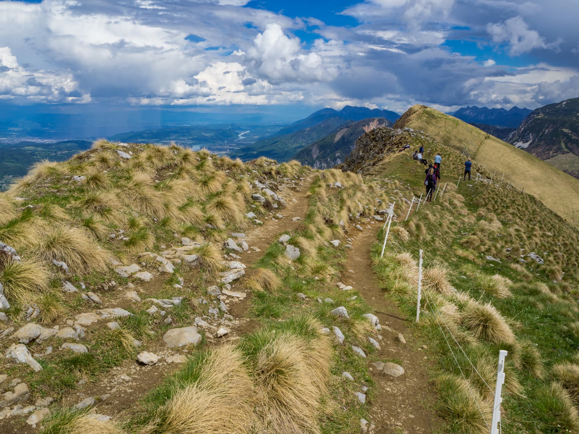 Hikers on a grassy ridge trail with distant mountains under a dramatic cloudy sky, Golica Ridge.