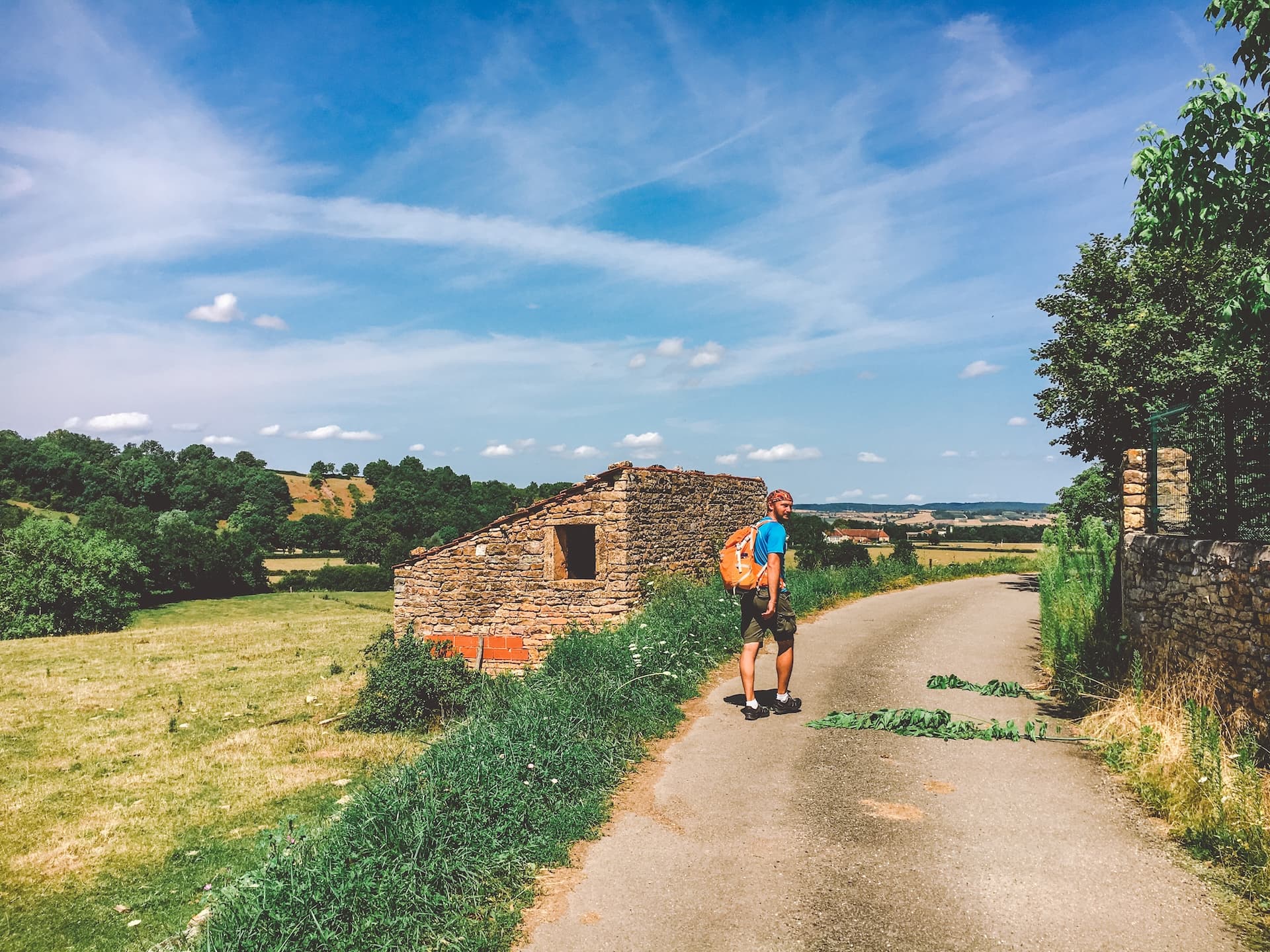 Hiker with orange backpack walking past stone ruin on rural road under blue sky.