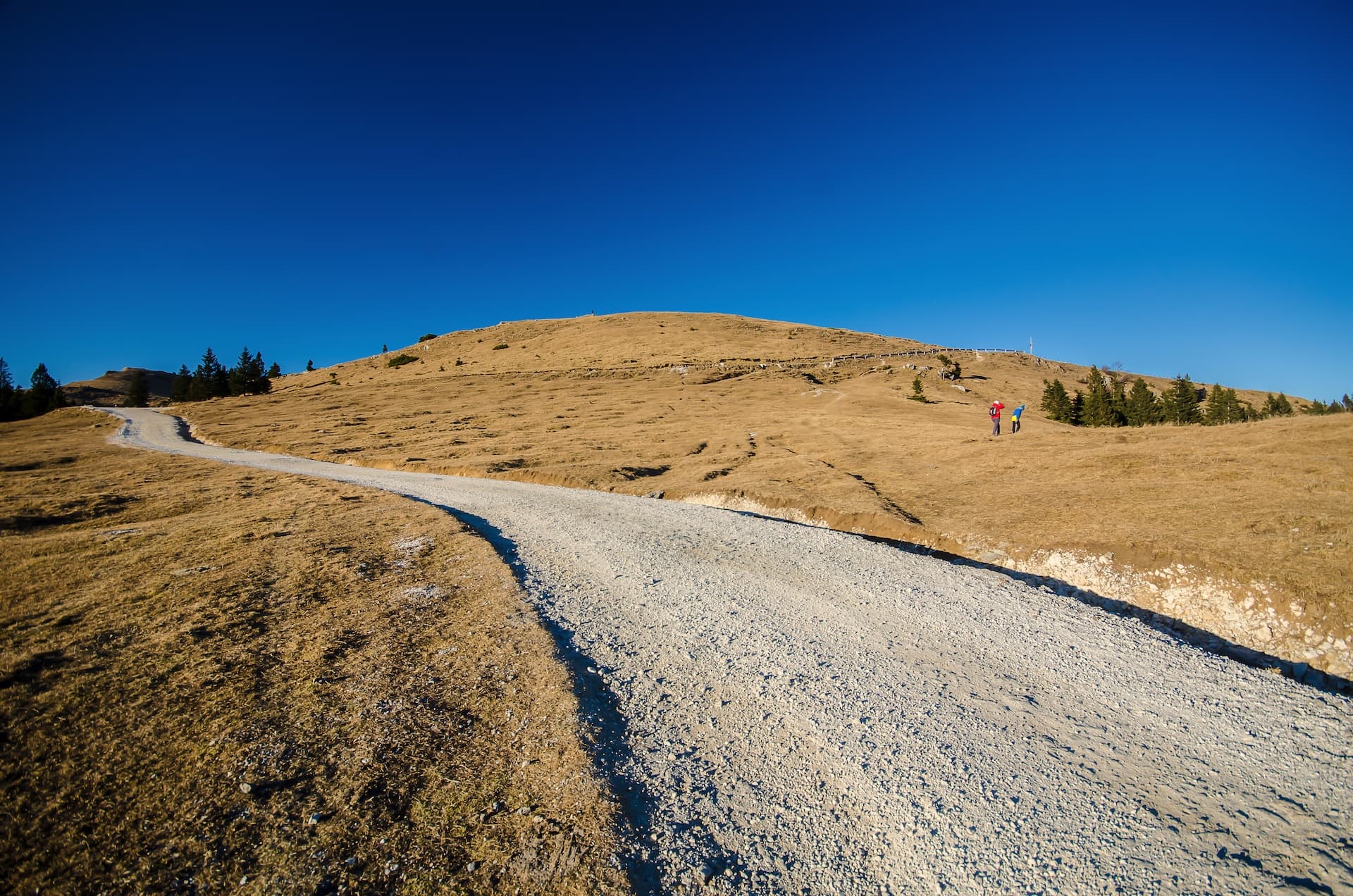 Hikers on gravel road ascending dry grassy hill under clear blue sky