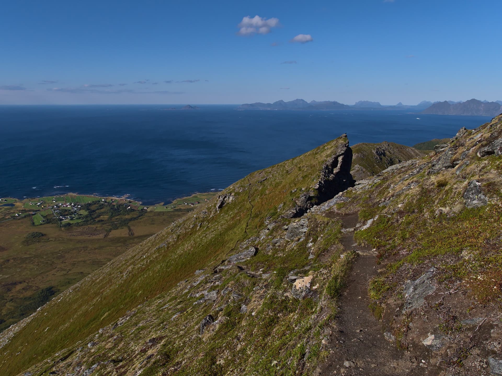 Hiking trail on grassy mountainside overlooking dark blue sea and distant islands under clear sky.