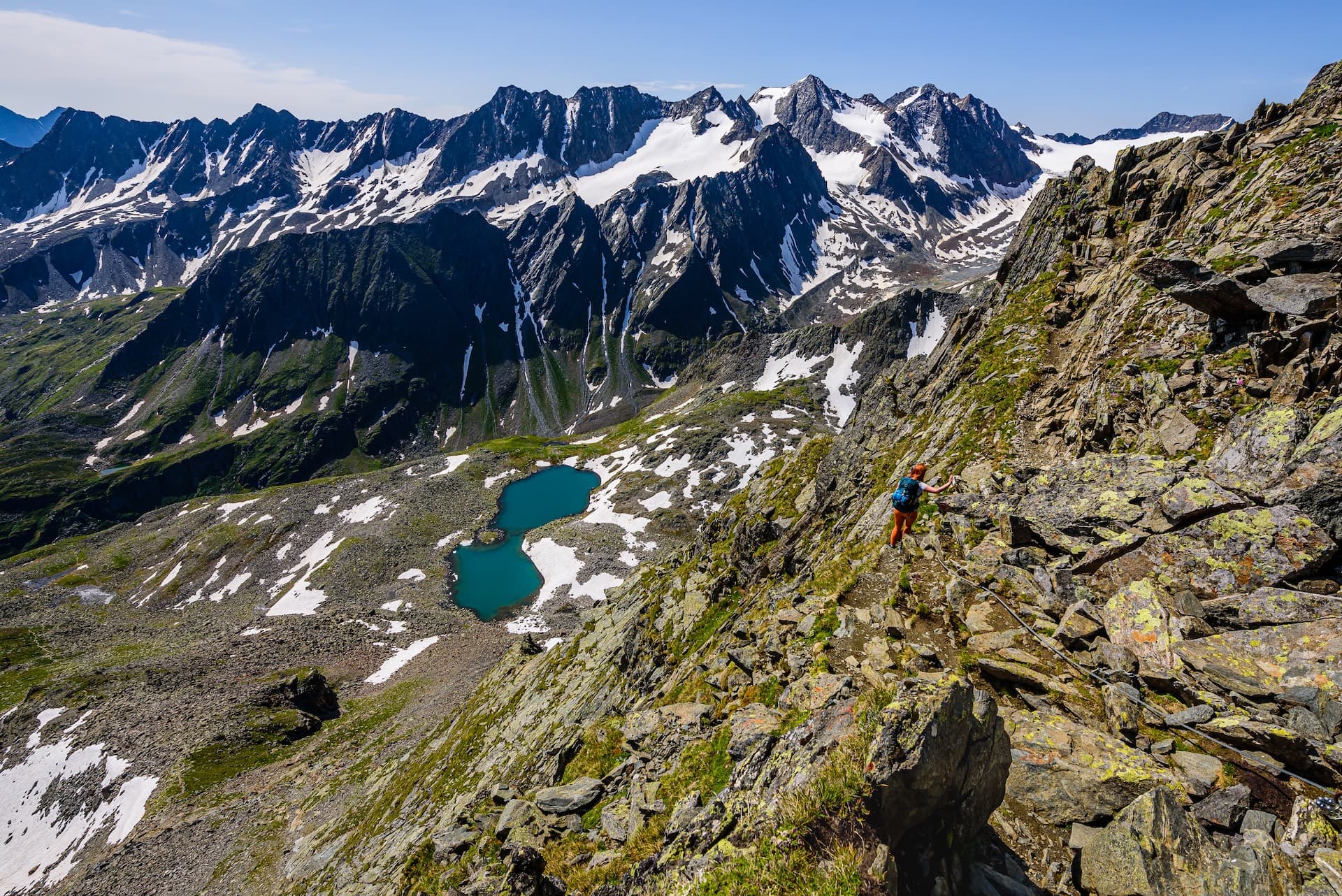Hiker ascending rocky alpine trail above turquoise glacial lakes with snow-capped mountains.