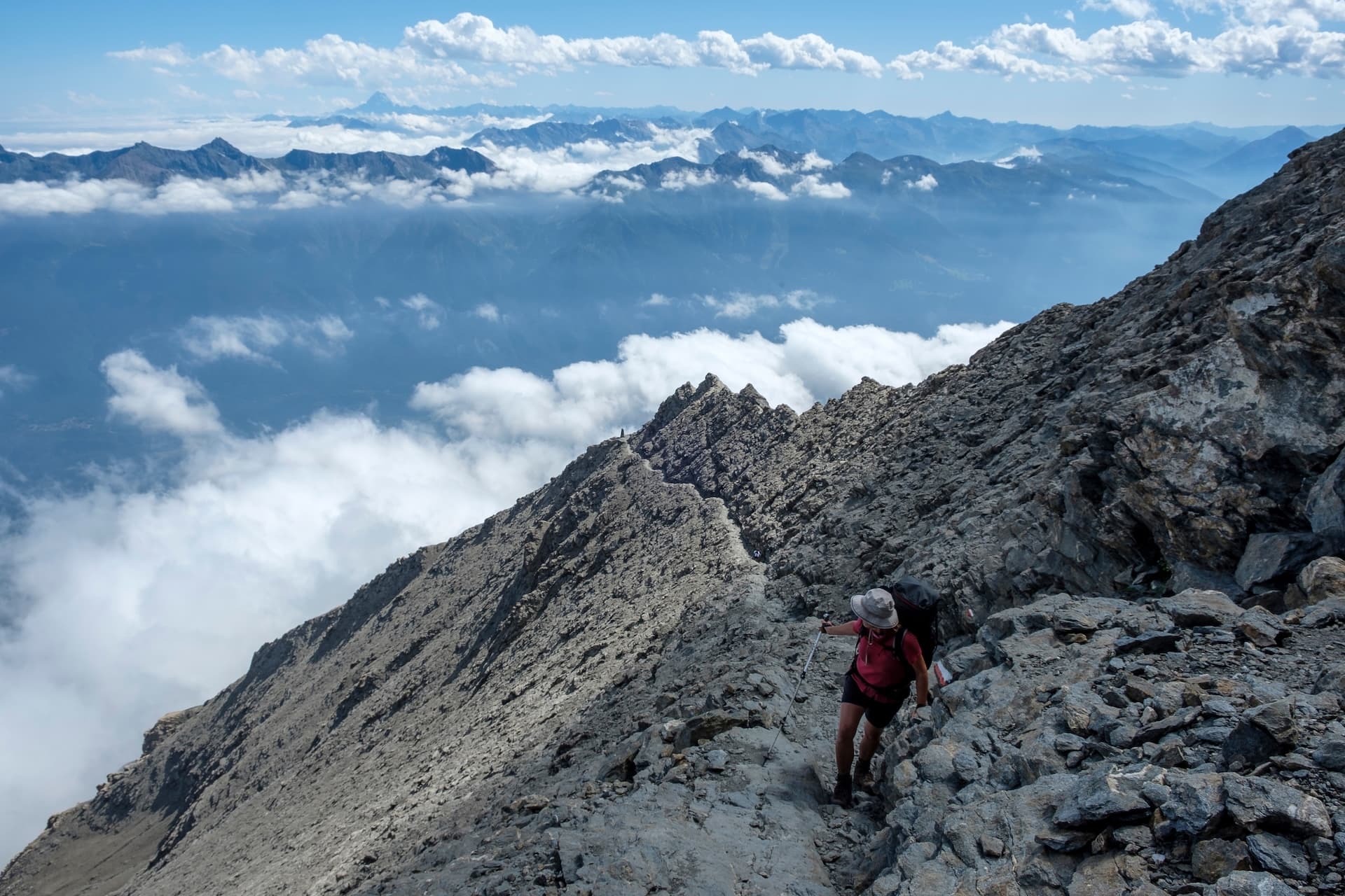 Hiker with backpack ascending steep rocky mountain ridge above clouds with distant peaks