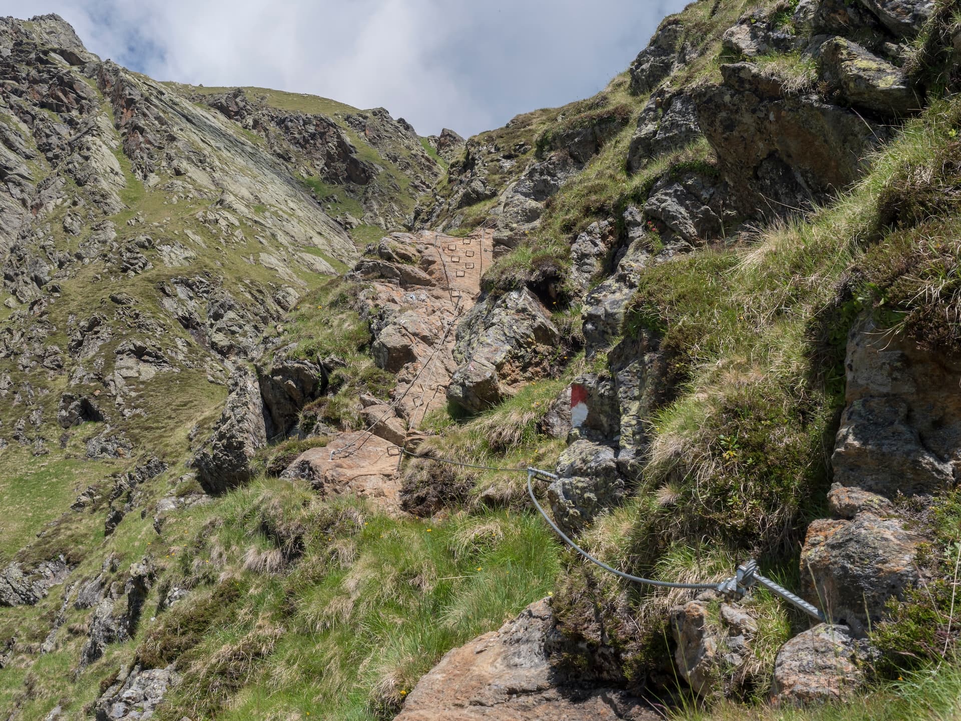 Via ferrata route ascending steep, rocky alpine terrain with green grass and cable protection.