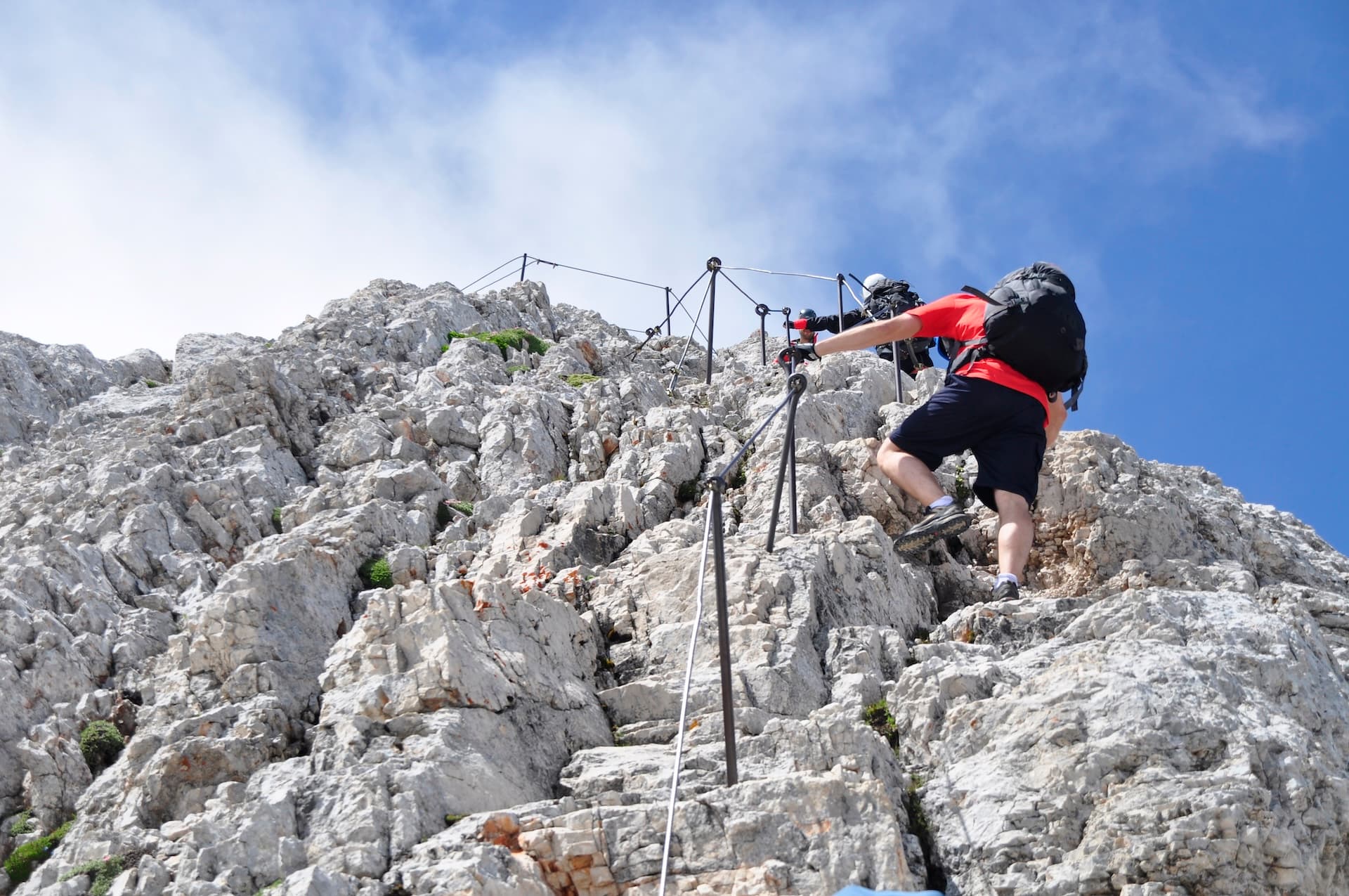 Hikers ascending steep, rocky mountain face using fixed cables under bright blue sky.