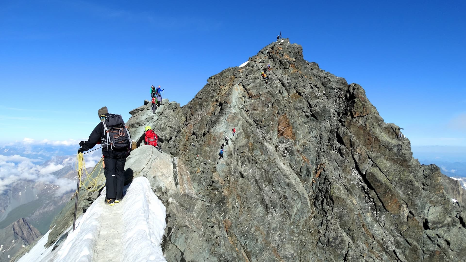 Mountaineers climbing steep, rocky peak with snow ridge under clear blue sky.