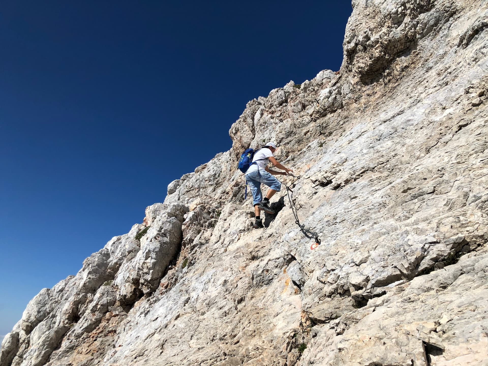 Climber ascending steep, rocky mountain face with via ferrata cables under clear blue sky.