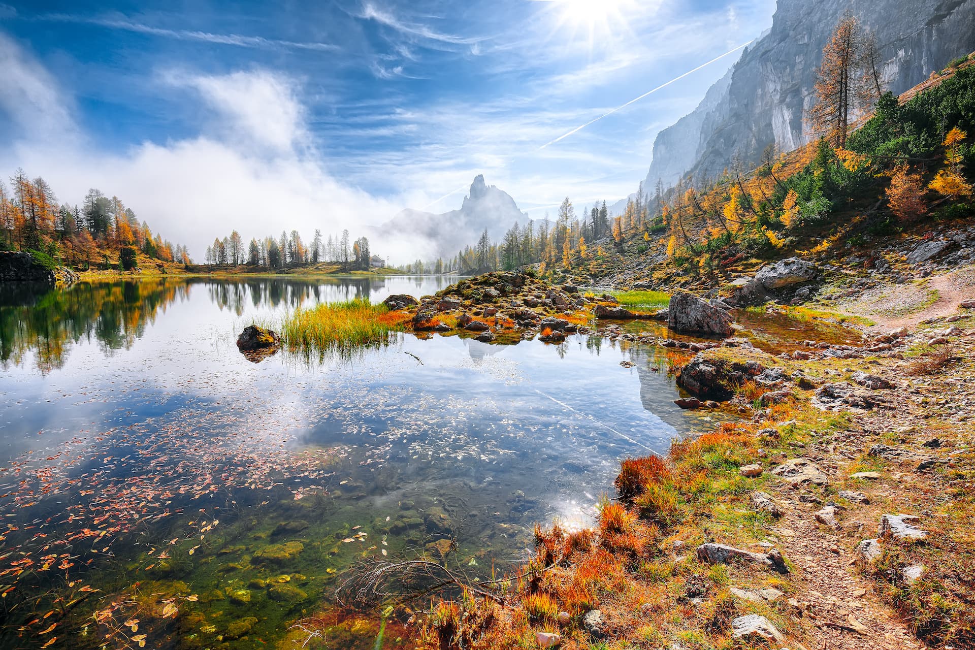 Mountain lake hiking trail with autumn foliage, clear water, and rocky shore under bright sun.