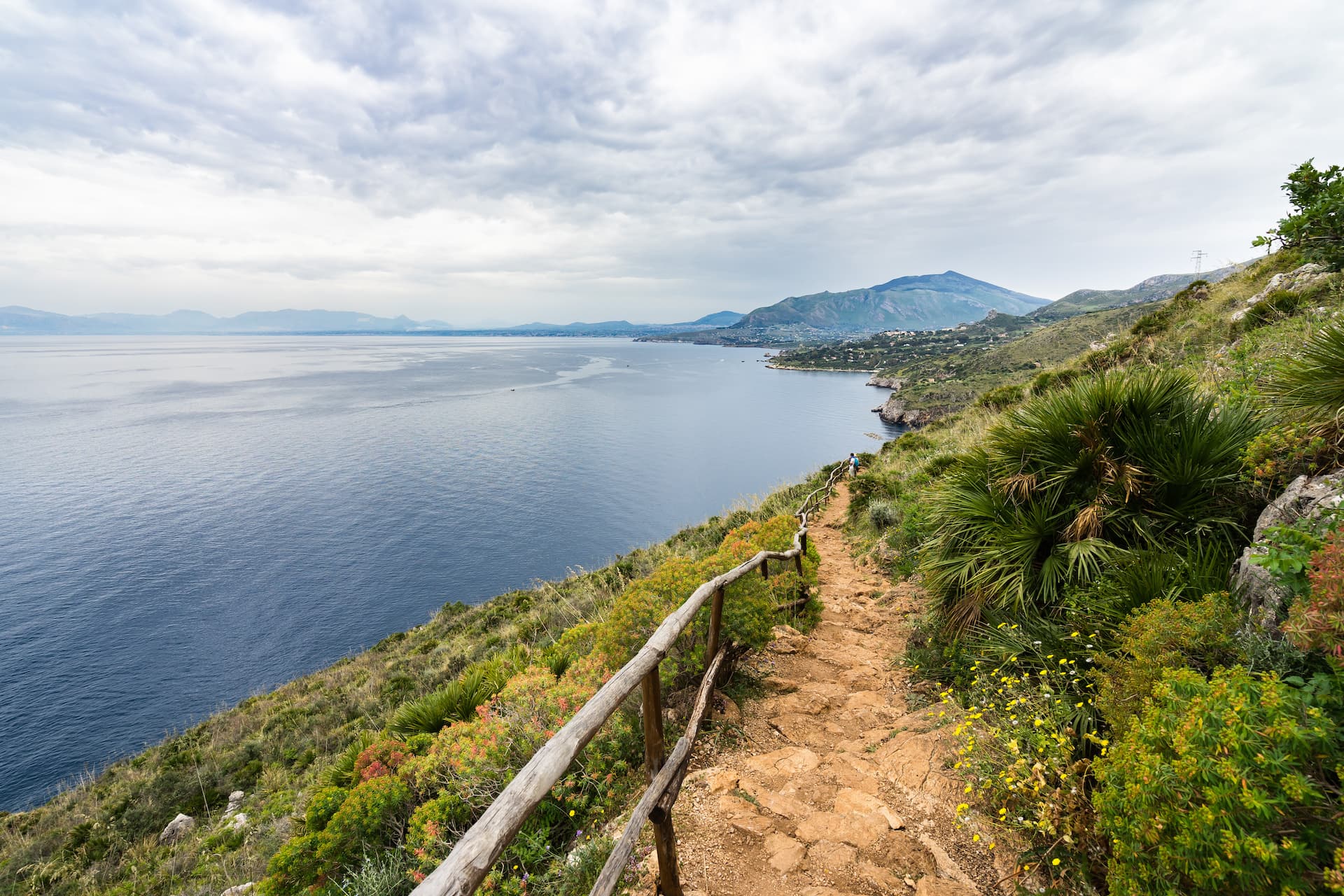 Coastal hiking trail overlooking blue sea, mountains, and green vegetation under cloudy sky.
