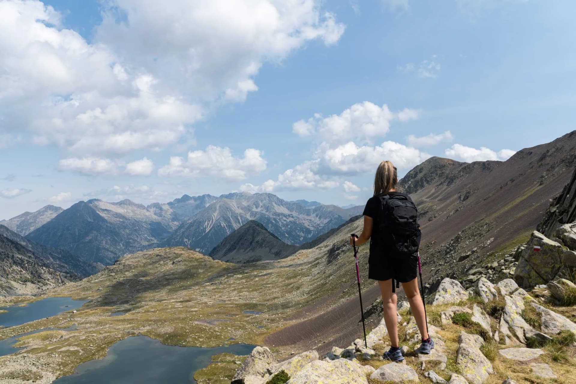 Hiker with poles overlooking mountain lakes and rugged peaks under a cloudy blue sky