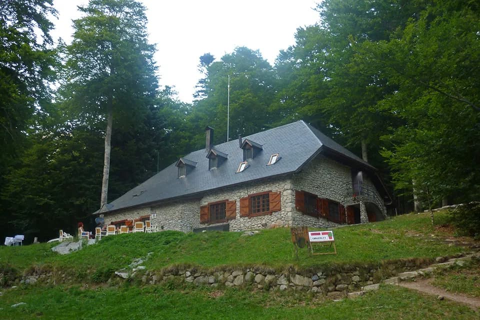 Stone mountain refuge Rifugio di Conangles surrounded by dense green forest with outdoor seating area.