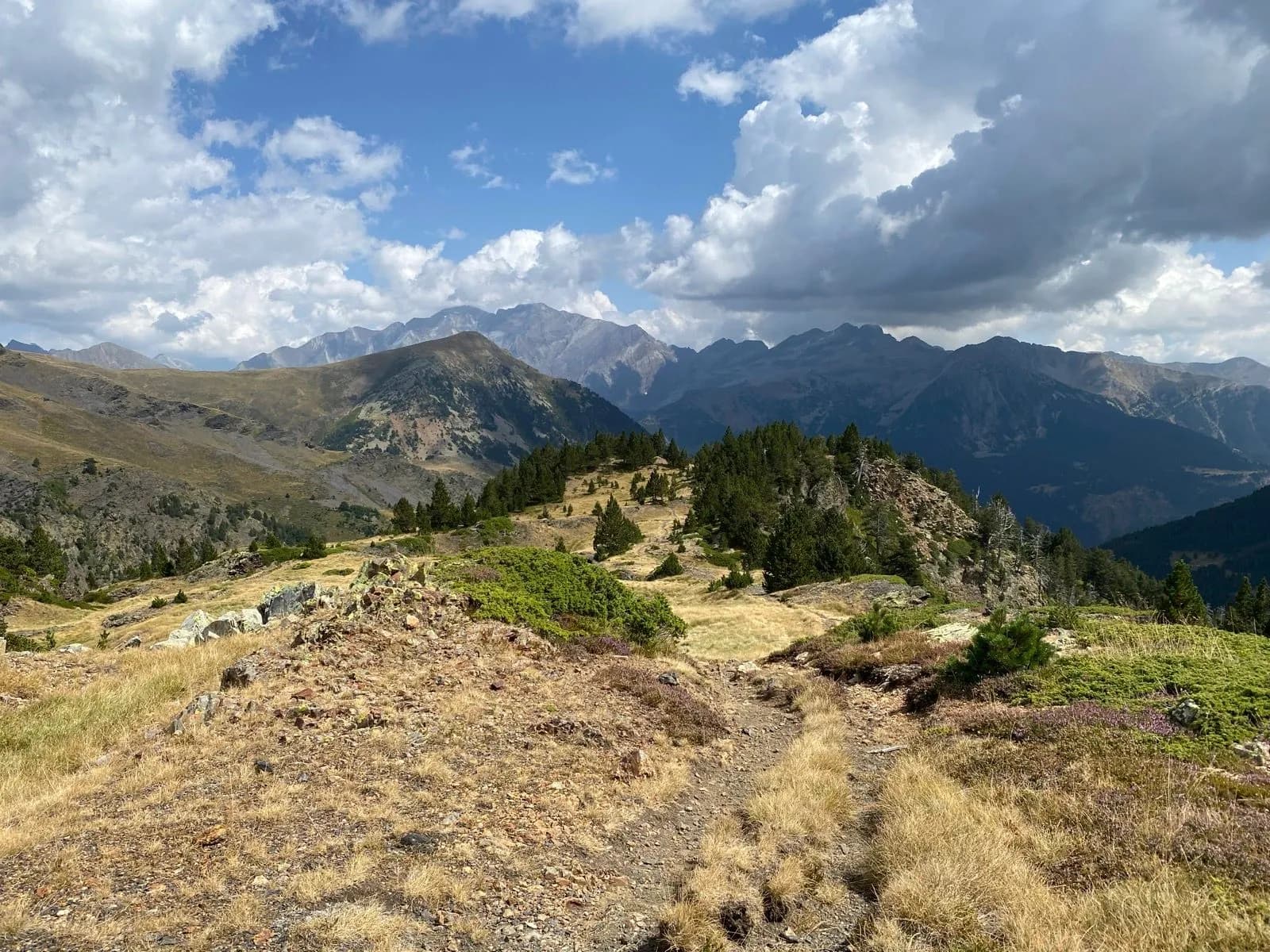 Hiking trail across dry alpine meadow toward distant, rugged mountains under cloudy sky.
