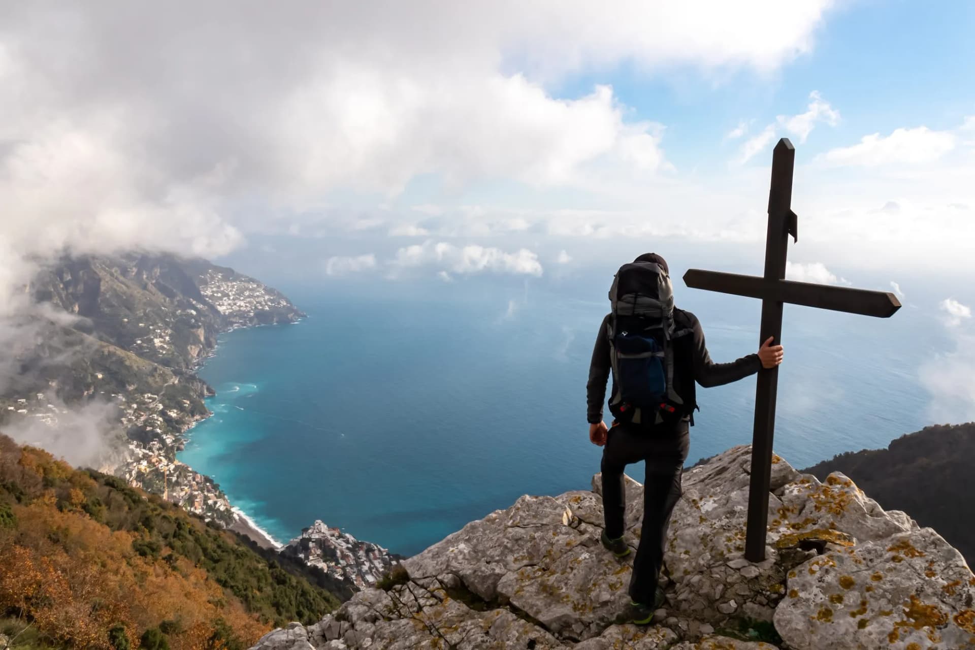 Active man with backpack at summit cross near Santa Maria del Castello. Scenic view on coastal town Positano. Magical hiking trail, Lattari Mountains, Apennines, Amalfi Coast, Campania, Italy, Europe