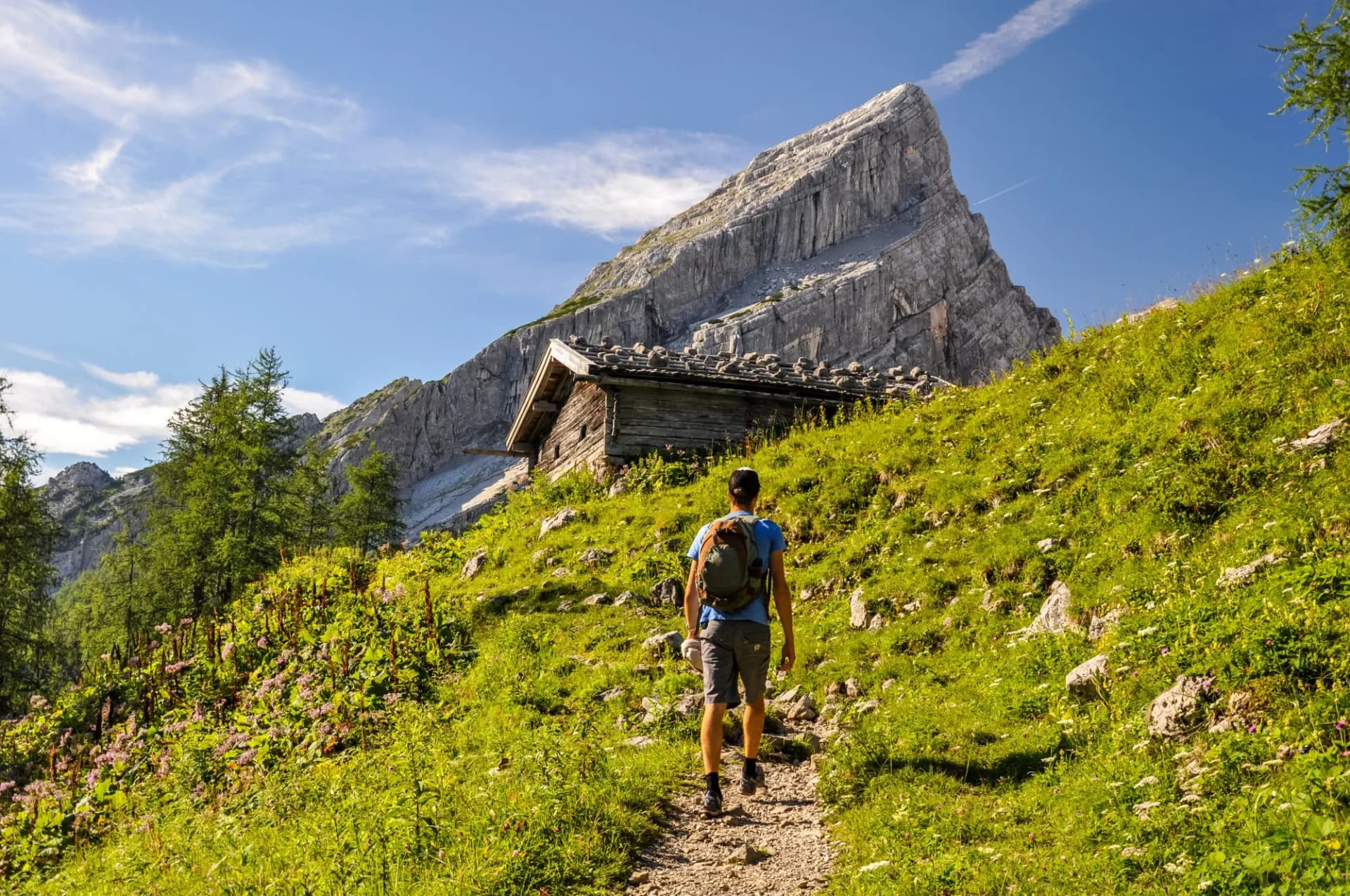 Young man hiking on a pathway up to Watzmann Mountain (2713m). In the background you can see a cabin and  "Kleiner Watzmann" (Little Watzmann, 2307m). Sunny day in the Bavarian Alps in Germany.