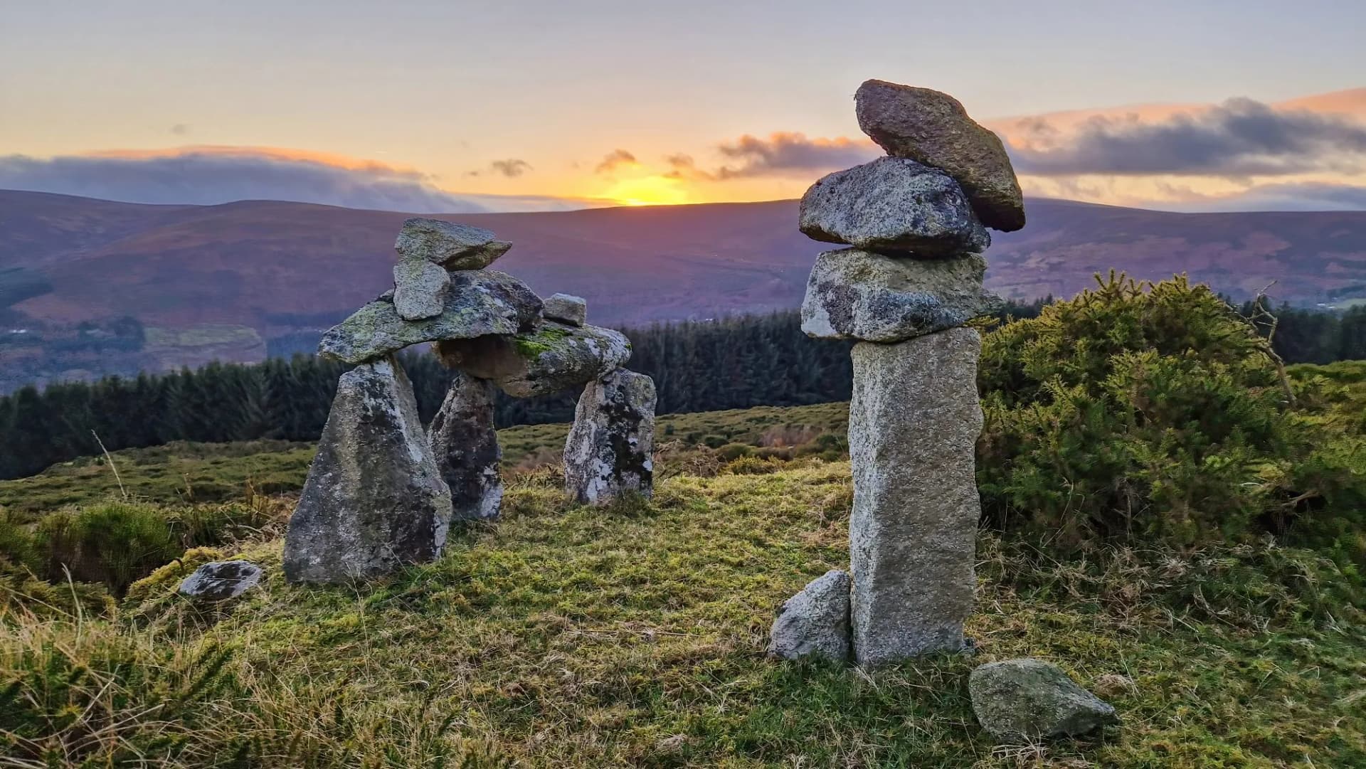 Stone cairn structures on grassy hillside at sunset overlooking rolling mountains at Glencree.