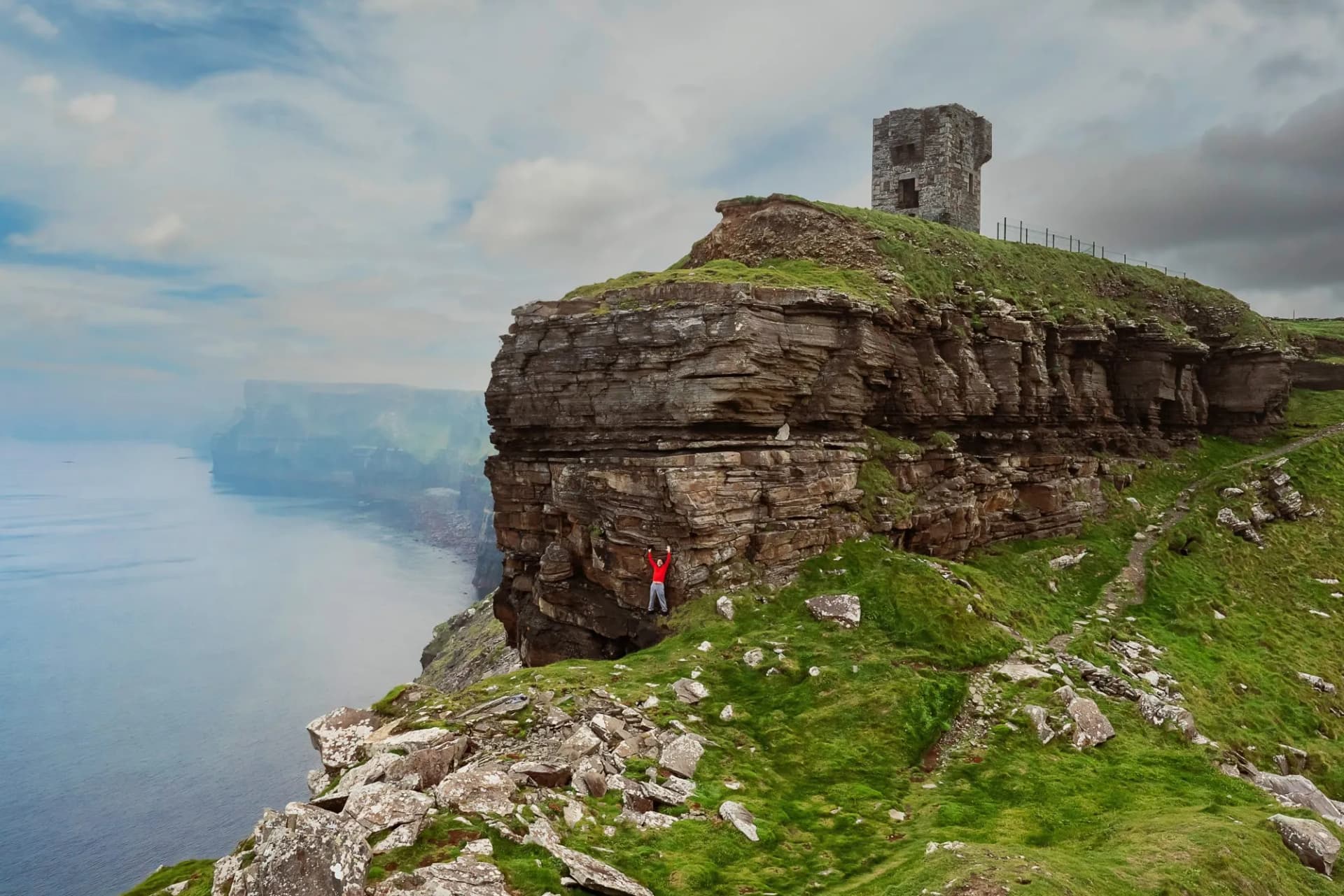 Male tourist standing on cliff with hands up in the air at Moher Tower at Hag's Head. Epic view of rock structure of Cliff of Moher, county Clare, Ireland. Cloudy sky.