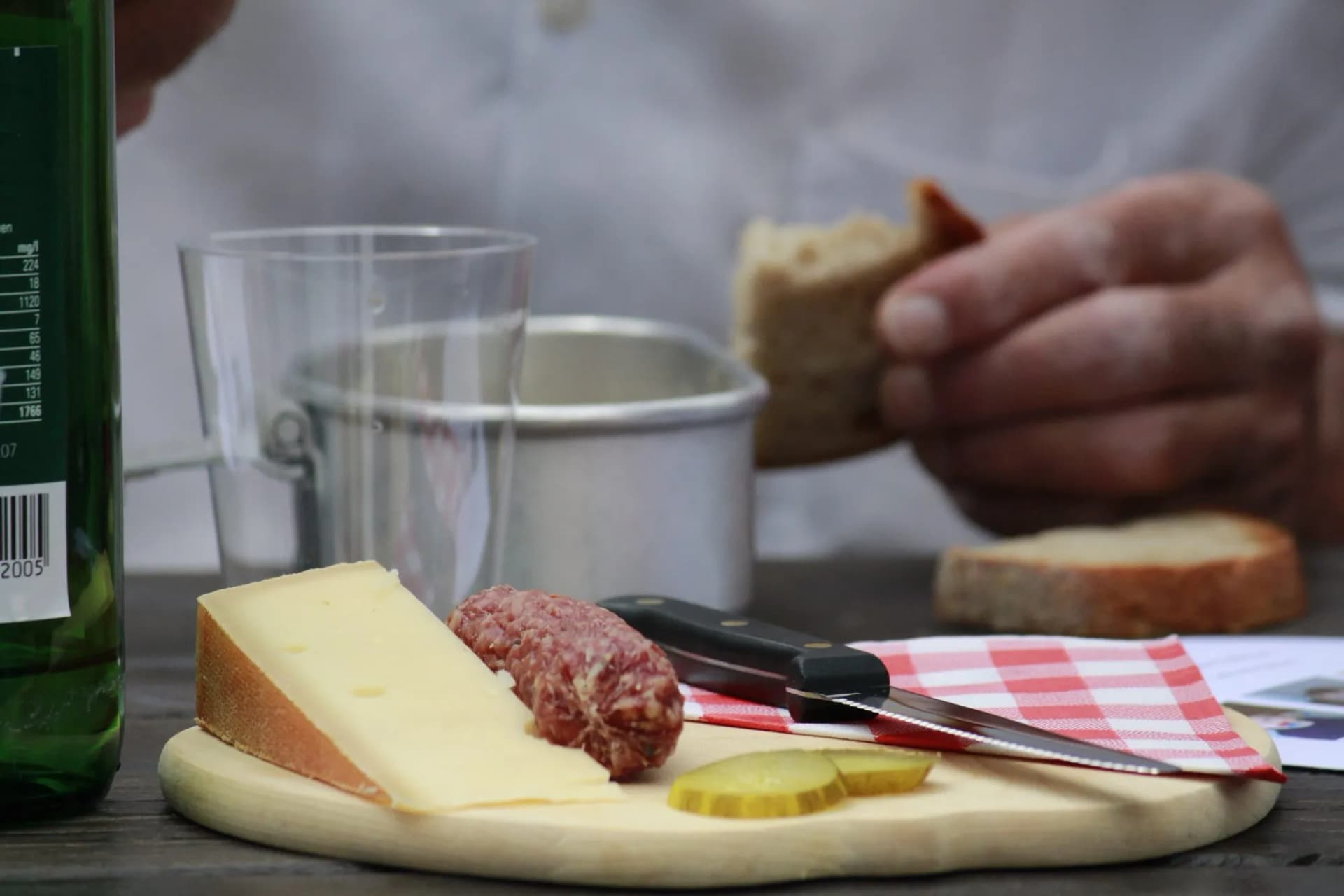 Cheese, salami, pickles, and bread on a cutting board with a knife and green bottle.