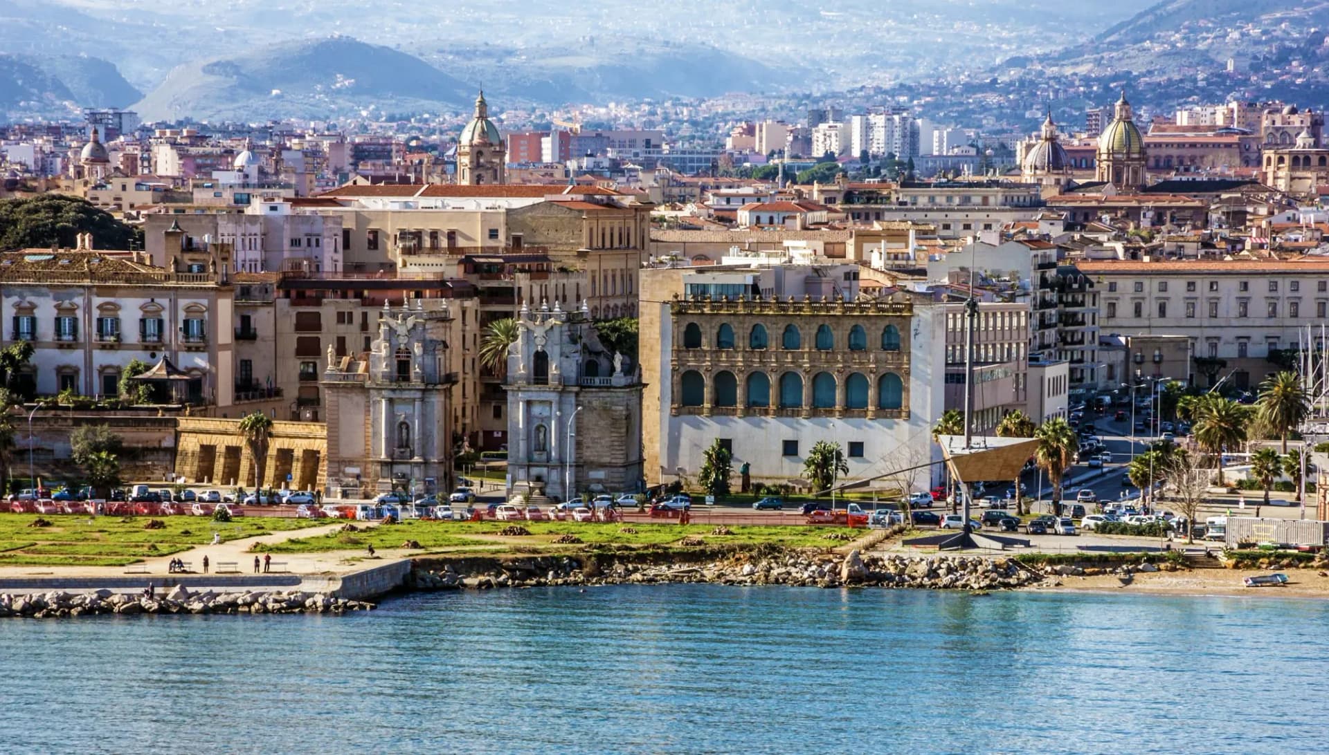 City skyline of Palermo with historic buildings and mountains in the background, viewed from the sea.