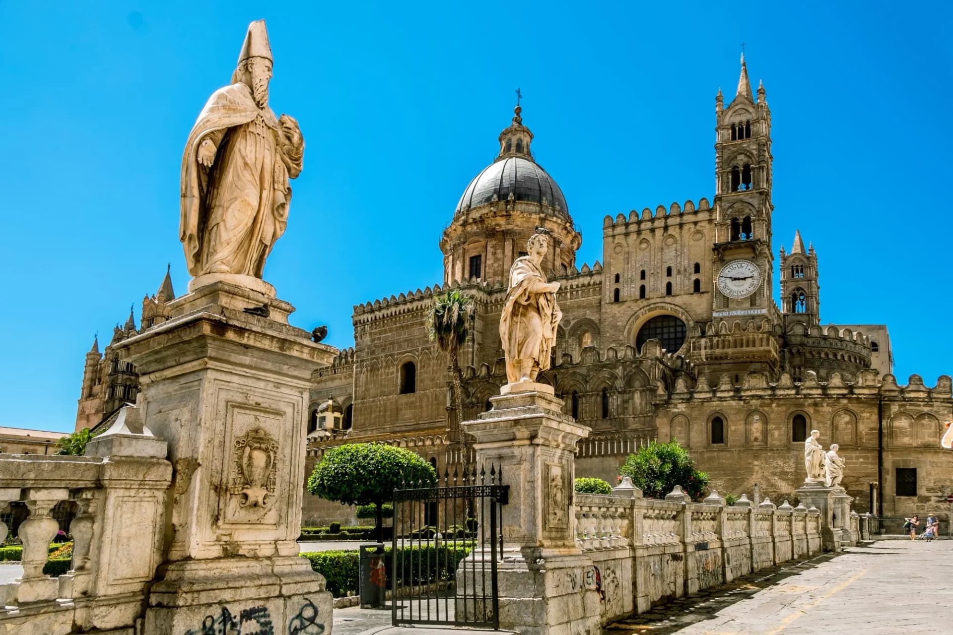Statues and historic Palermo Cathedral with dome and clock tower under bright blue sky.
