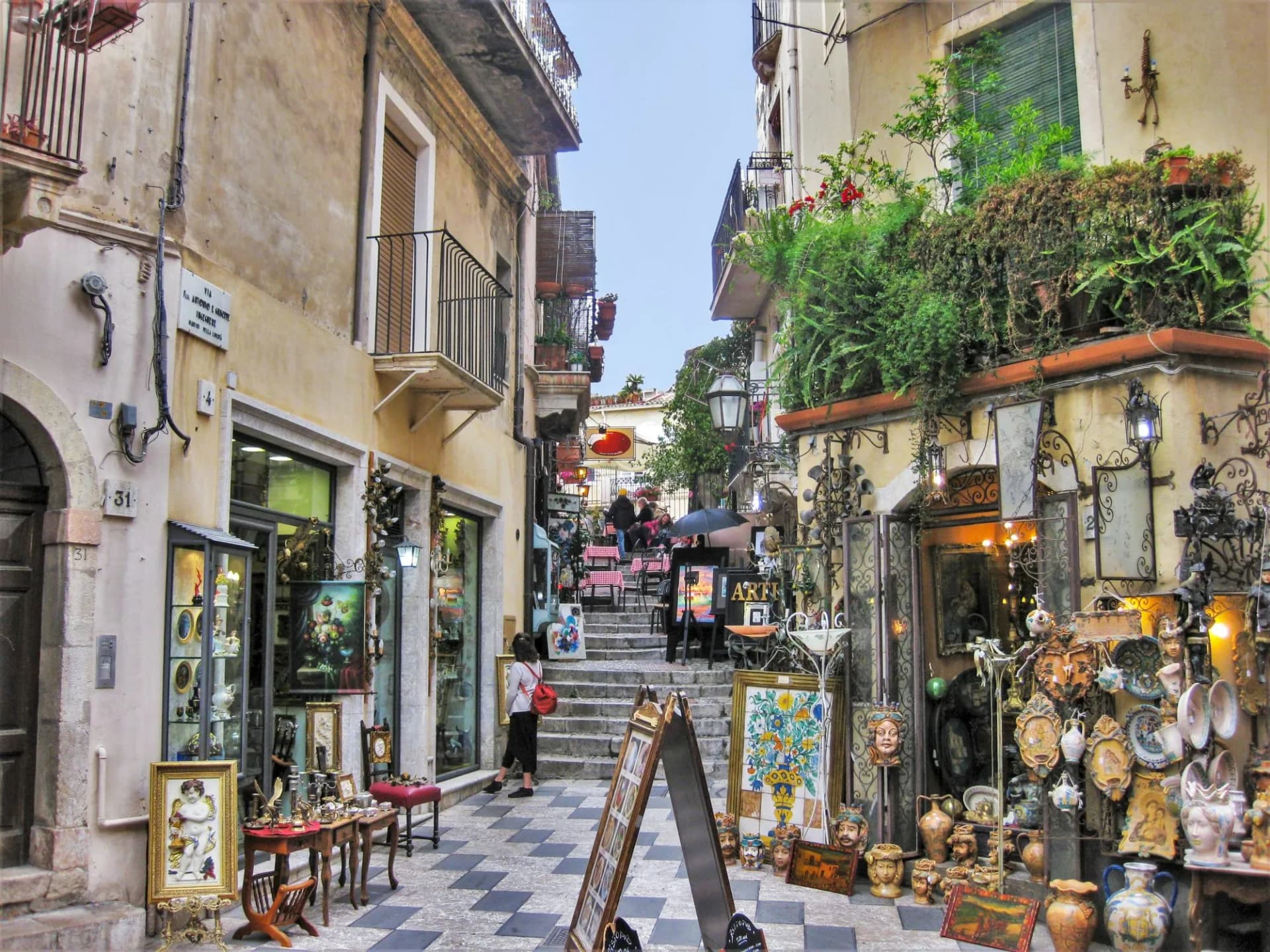 Colorful street in Taormina, Italy, with shops displaying art and ceramics leading to stairs.
