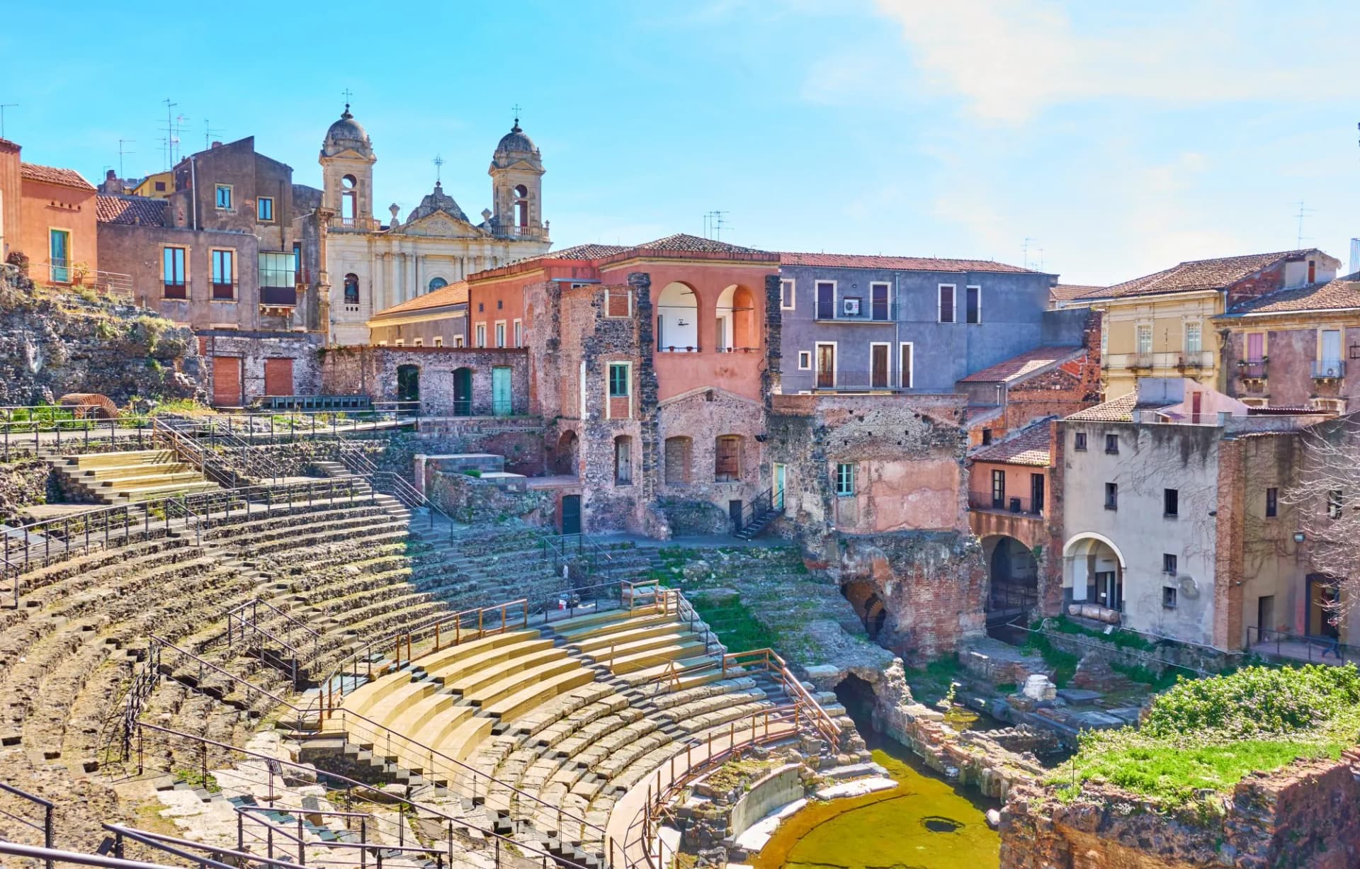 Ancient Roman theatre ruins with tiered seating and historic buildings in Catania, Sicily.