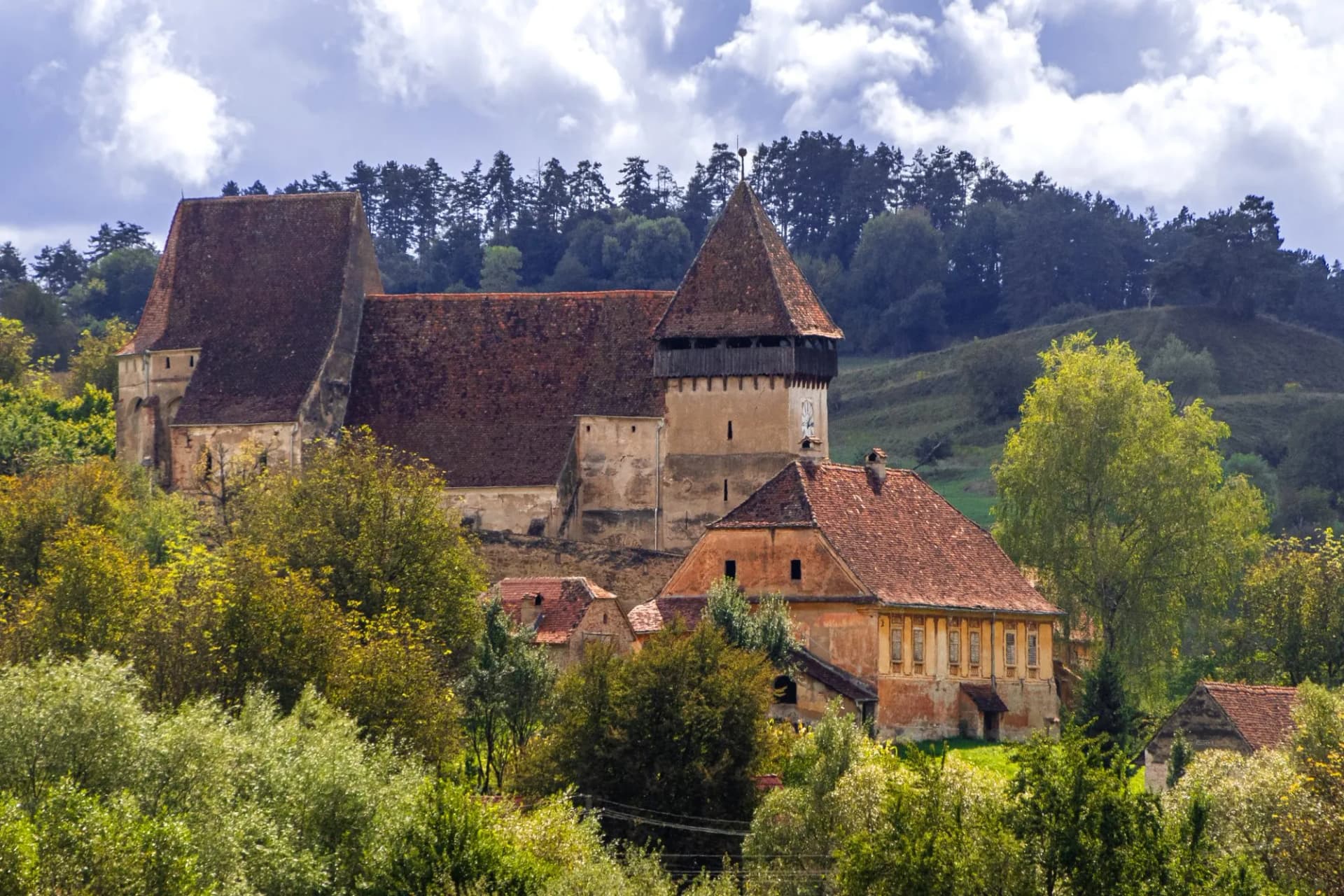 Fortified church with red tile roof and tower surrounded by green hills and trees under cloudy sky.