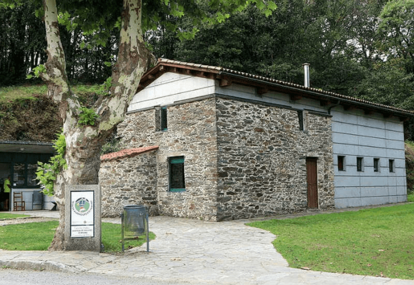 Stone albergue building with modern extension, large tree, and path in a wooded area.