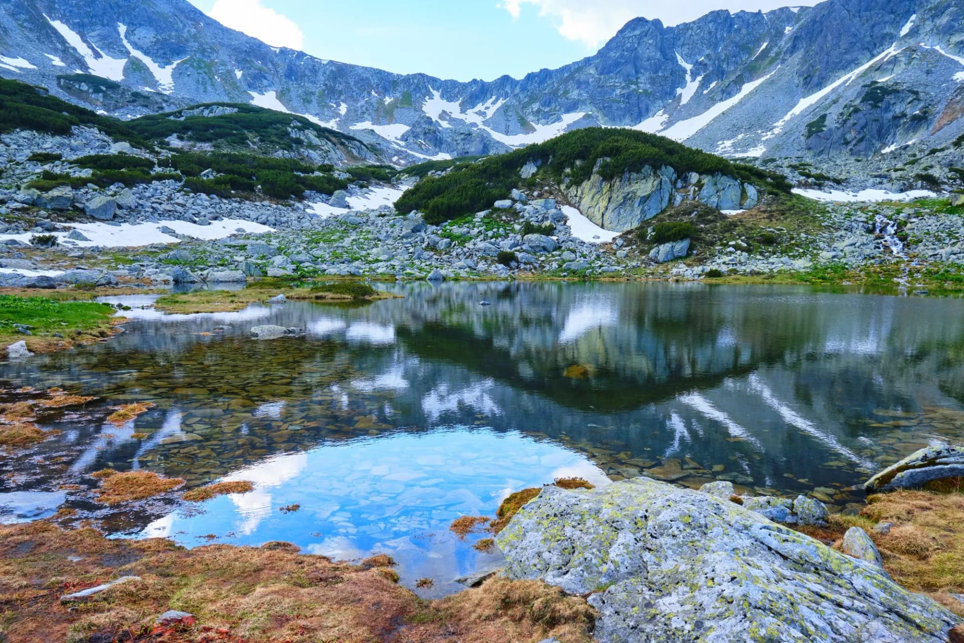Perfect reflection in a mountain lake (Pietrele Lake, Retezat mountains), with brown moss at shore, rocks in clear water, tall mountain ridge (Curmatura Bucurei) in the back, blue sky, spots of snow.