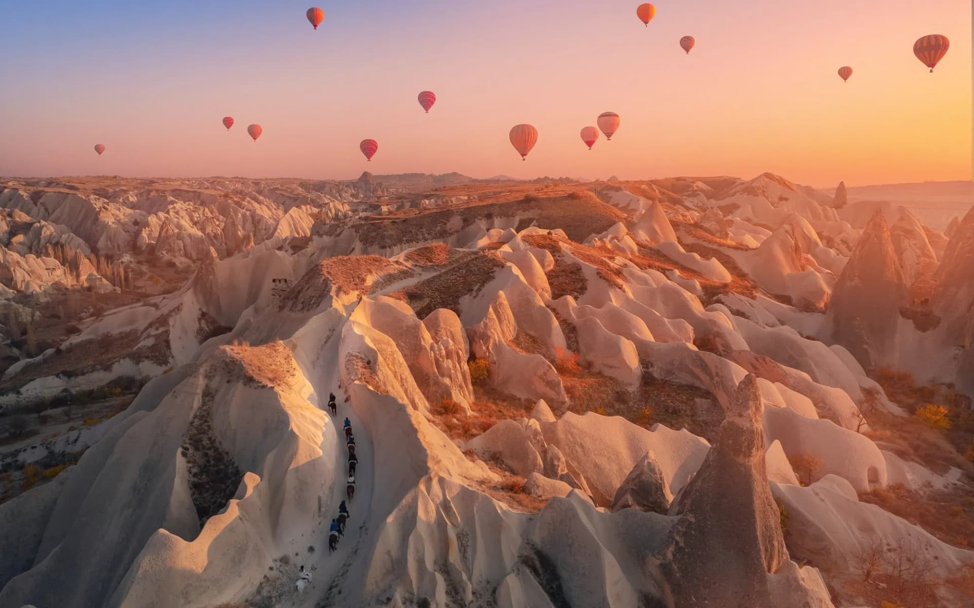 Hot air balloons over Rose Valley at sunset with people horseback riding through rock formations.