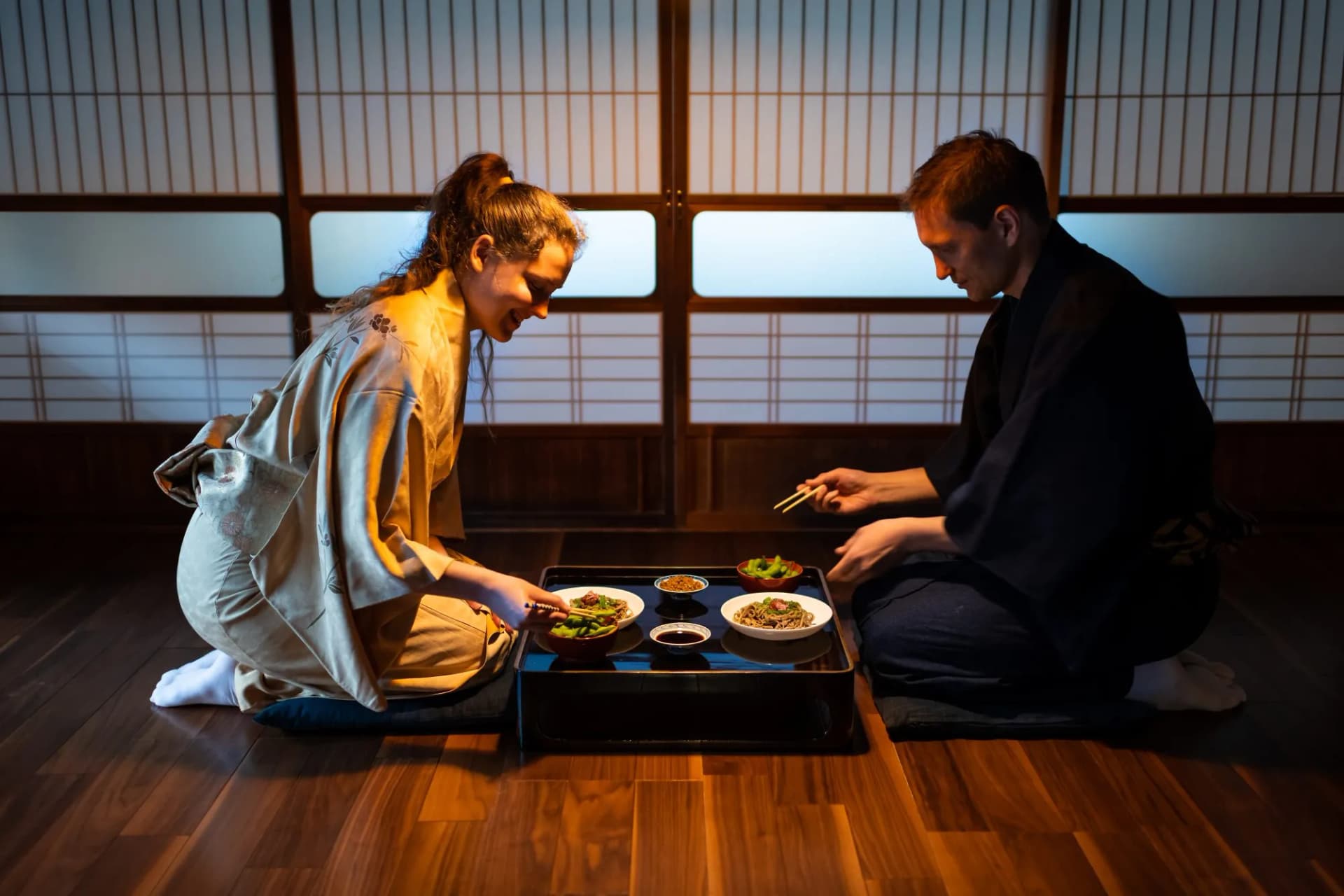 Young couple seiza sitting on floor pillows with Japanese ryokan kaiseki dishes, eating soba noodles, natto, edamame and soy sauce on table by sliding paper doors