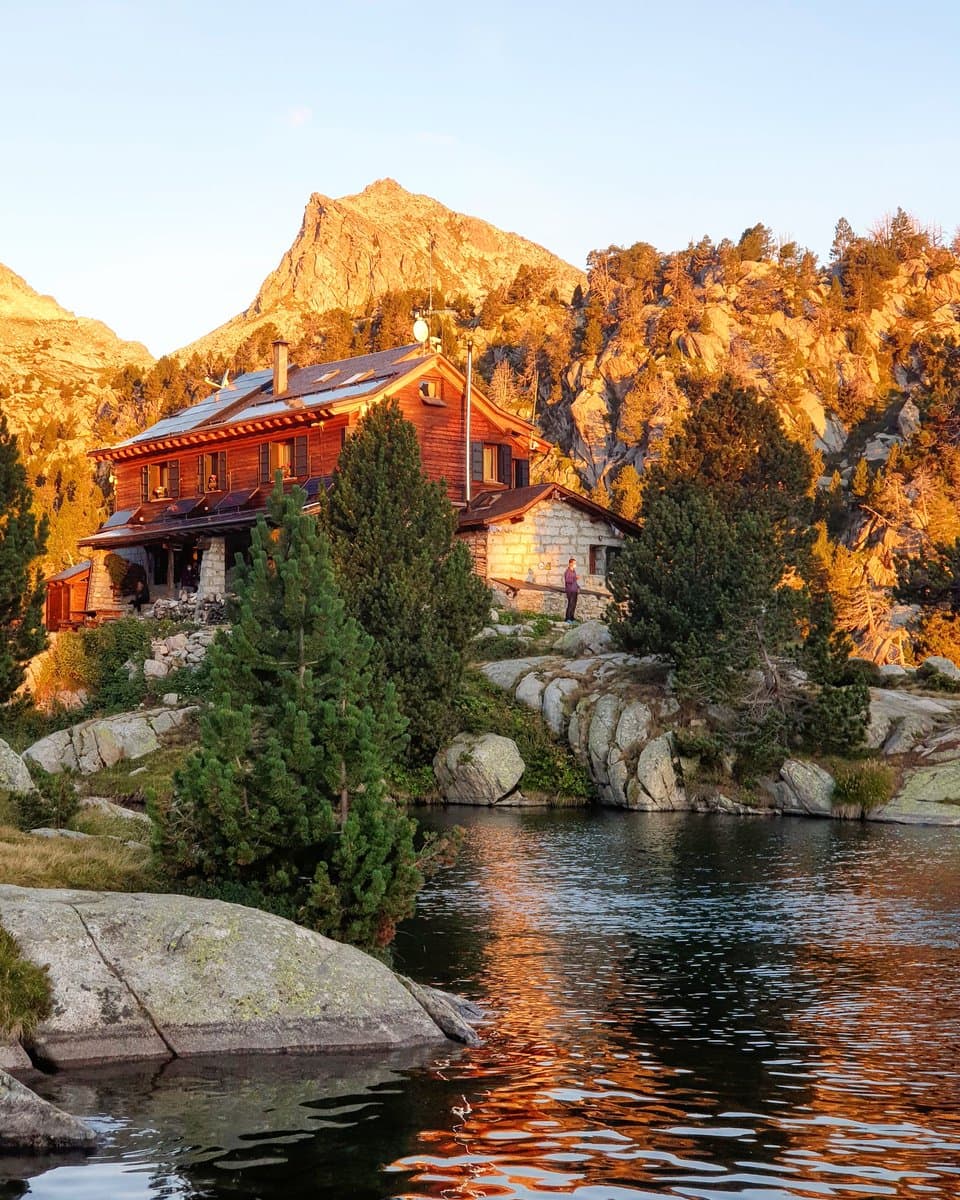 Wooden mountain lodge by a lake with rocky peaks at sunset.