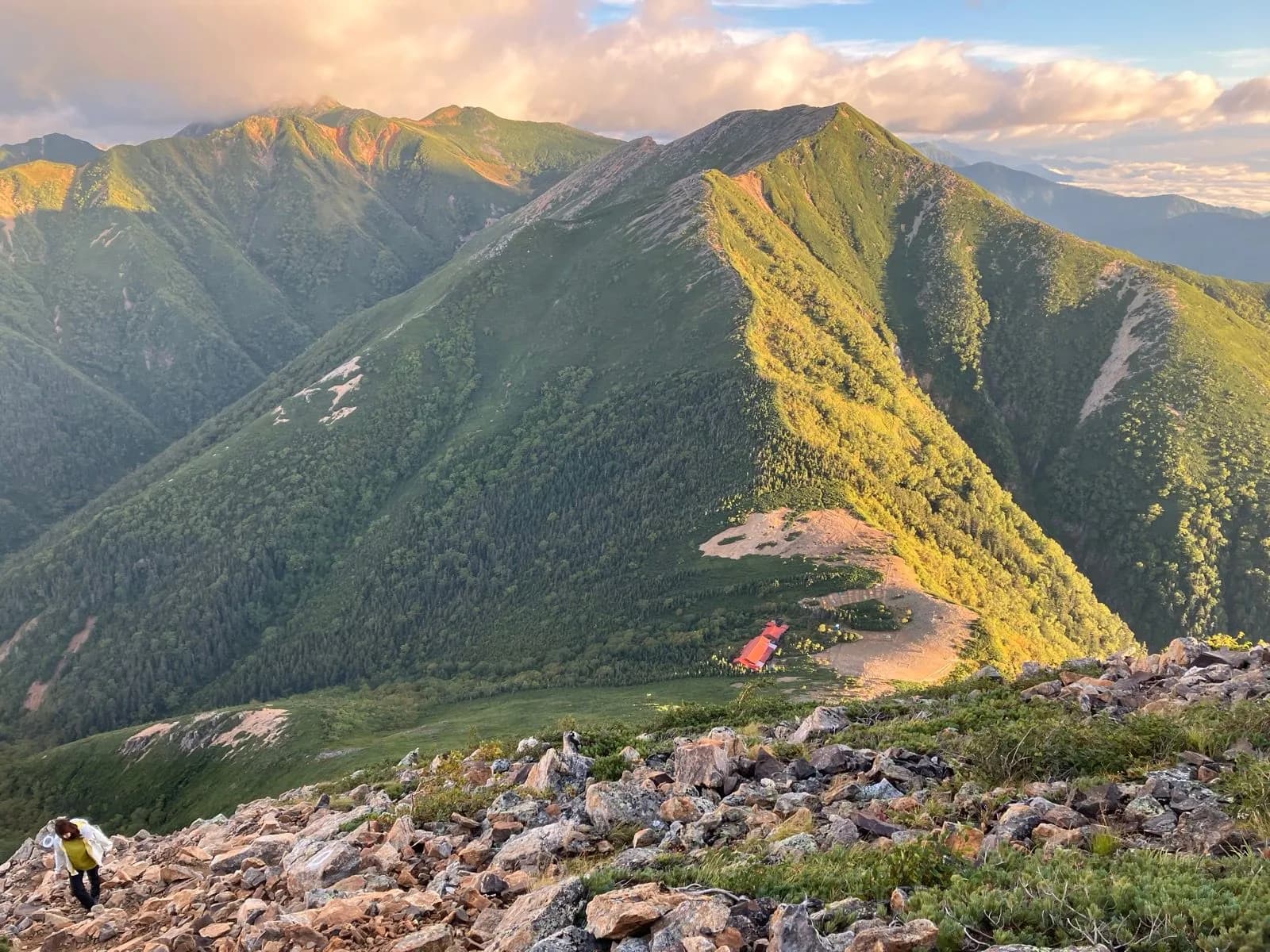 Hikers on rocky ridge with green mountains and orange-roofed hut, Chogatake and Daitenso.