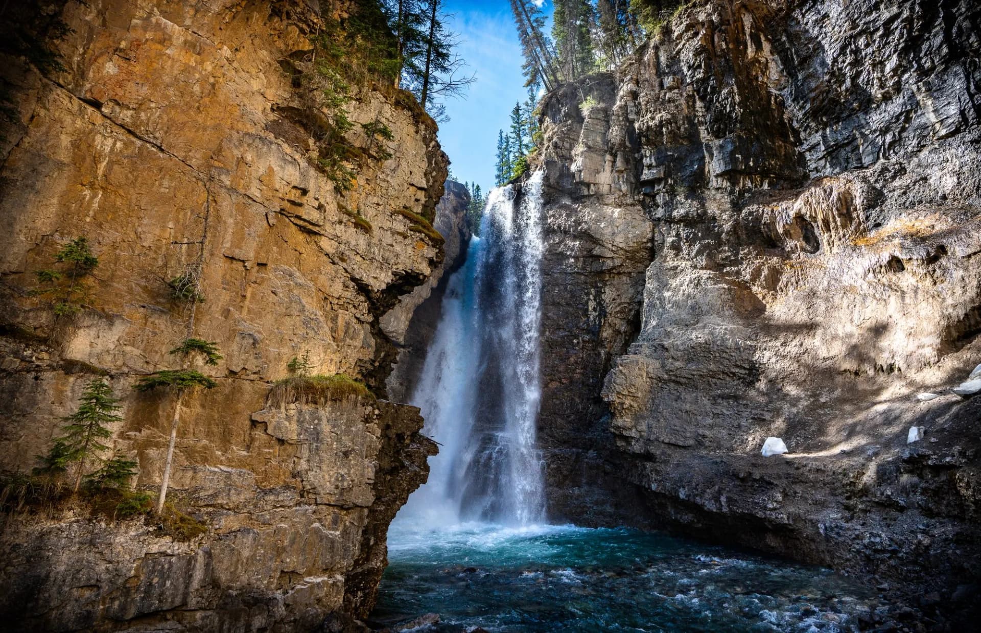 Waterfall cascading between tall, rocky canyon walls with evergreen trees under blue sky at Johnston Canyon.