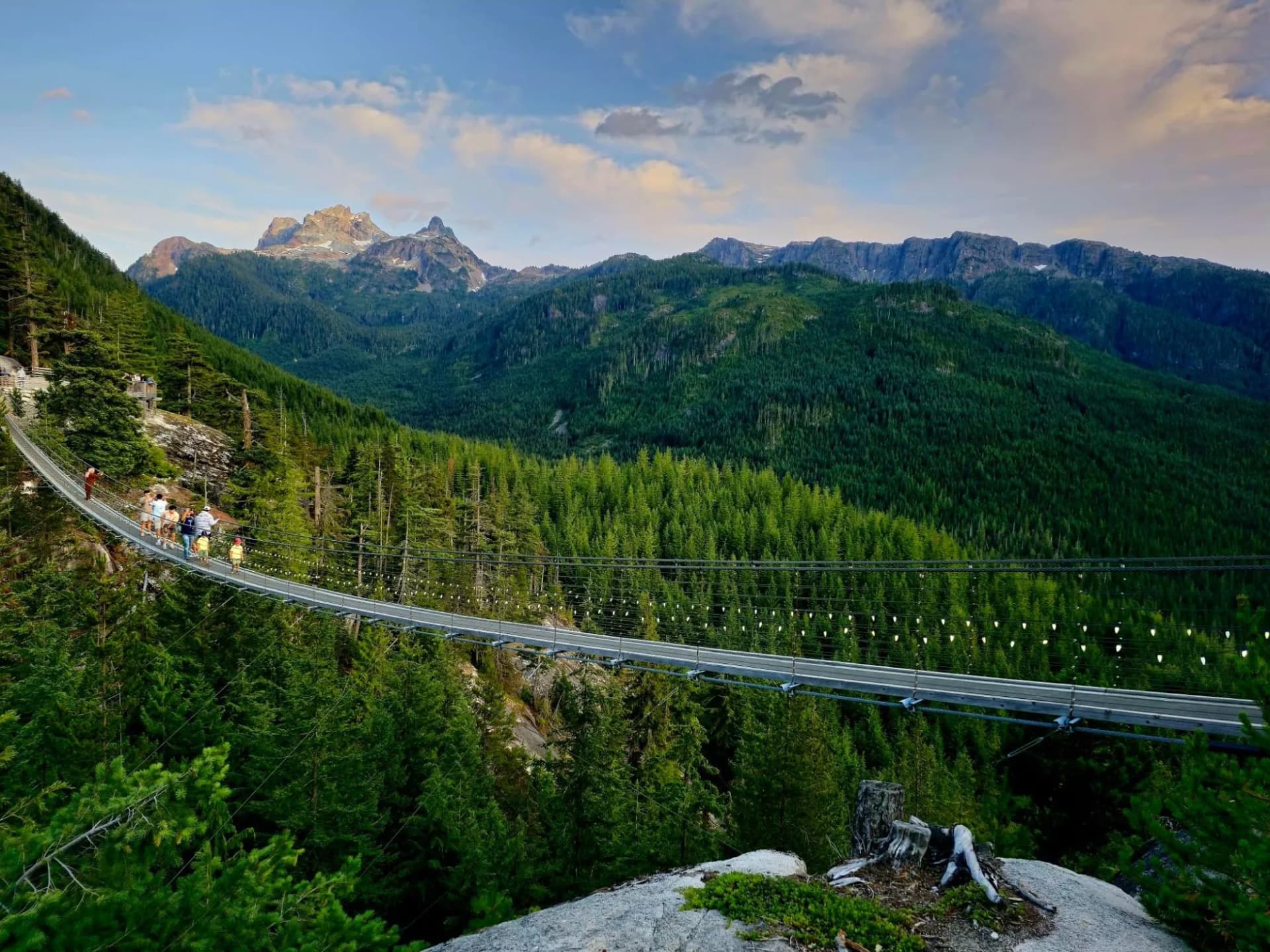 Suspension bridge over dense evergreen forest with rugged mountains under a cloudy sky