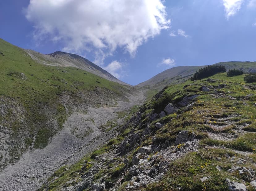 Hiking trail leading up a grassy alpine mountain slope with rocky terrain under a blue sky.
